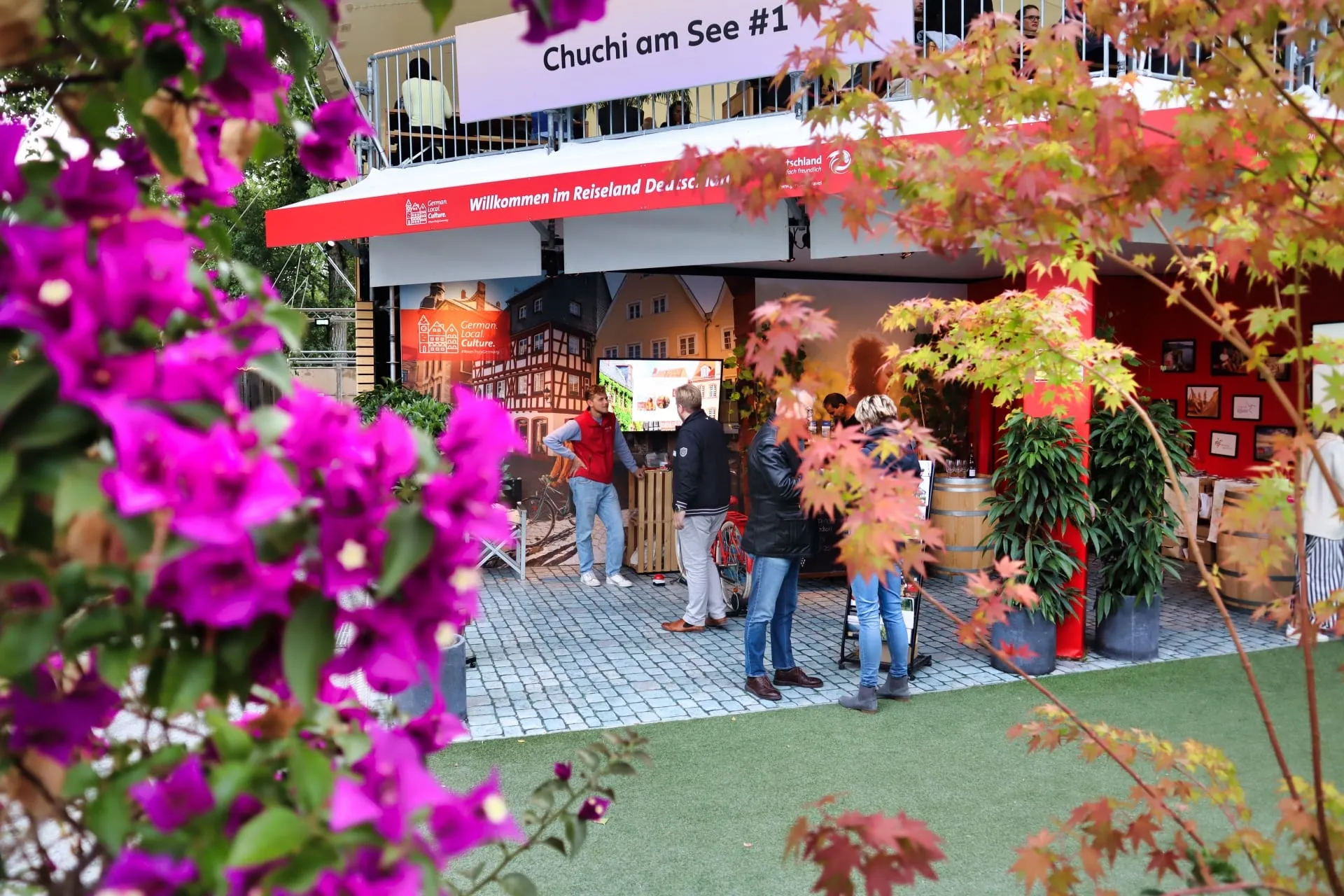 Red DZT stand with a photo wall, wooden barrels and visitors beneath the balcony of «Chuchi am See»