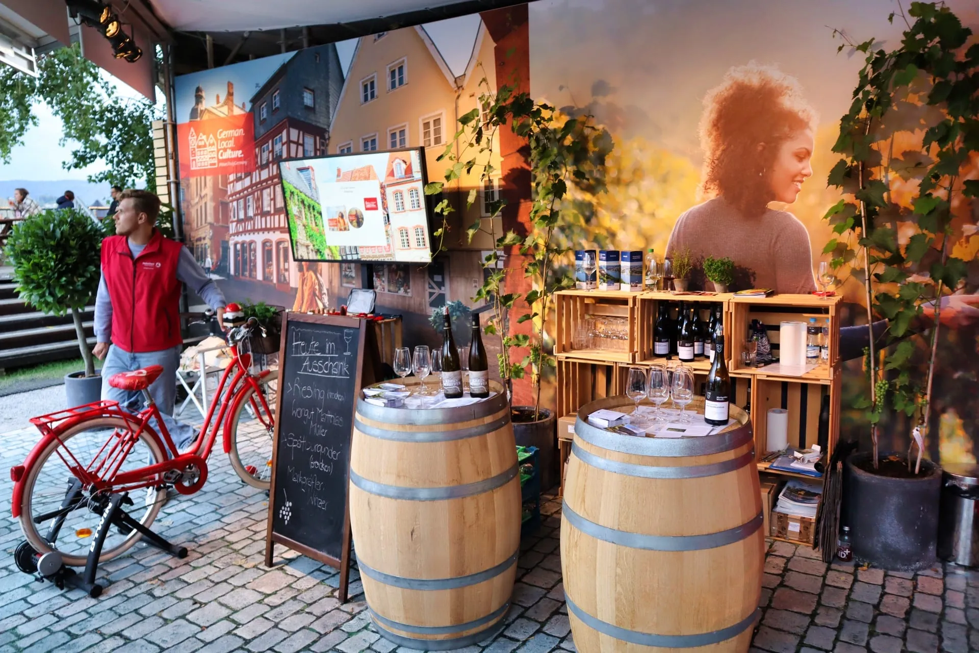 Rotes Velo auf Rollentrainer, Kreidetafel und Weinfässer mit Gläsern vor der Fotowand des DZT-Stands