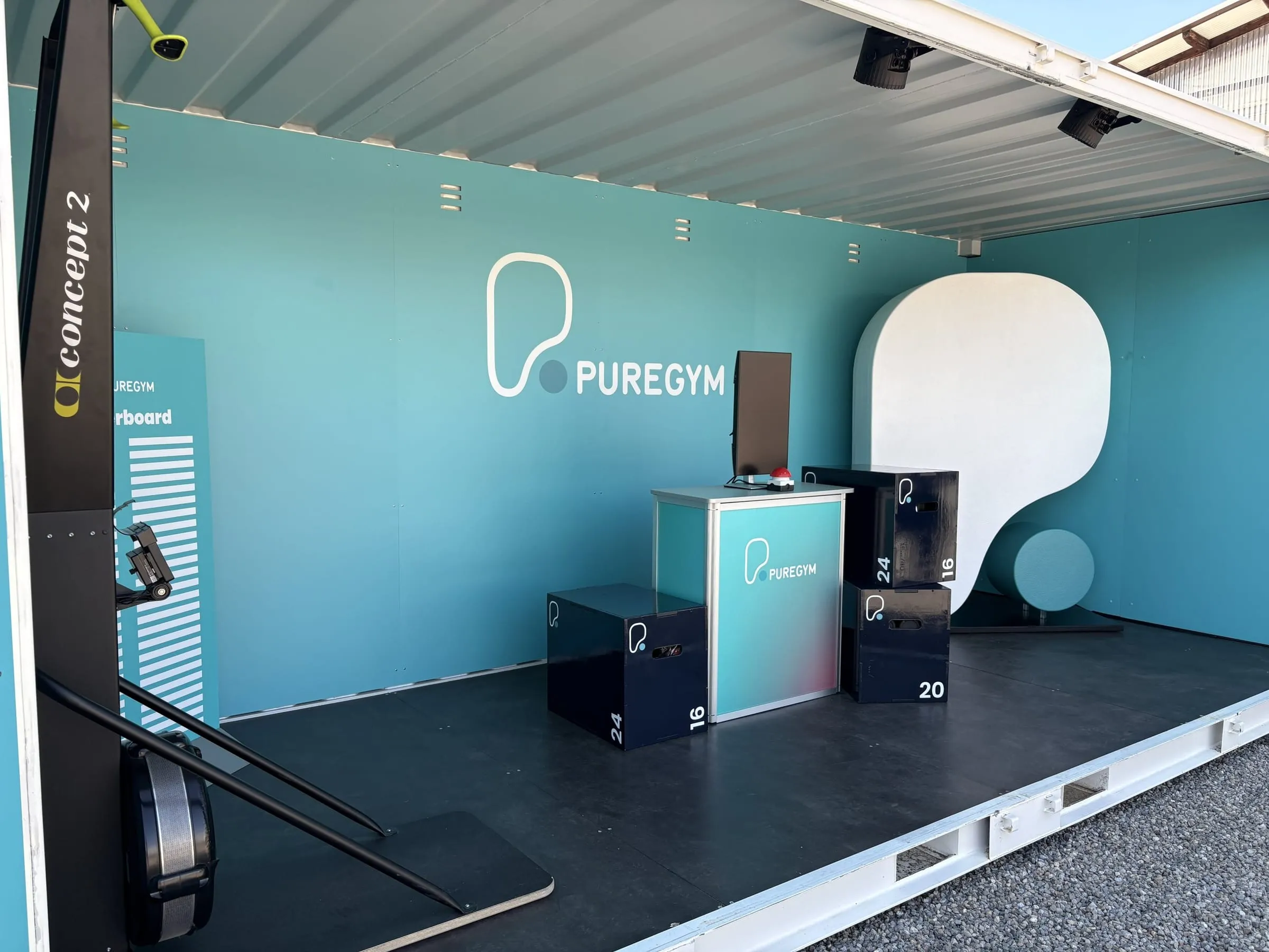 Blick in den türkisen Container mit PureGym-Logo, Theke mit Screen und rotem Buzzer, schwarzen Plyo-Boxen und weisser Logoform