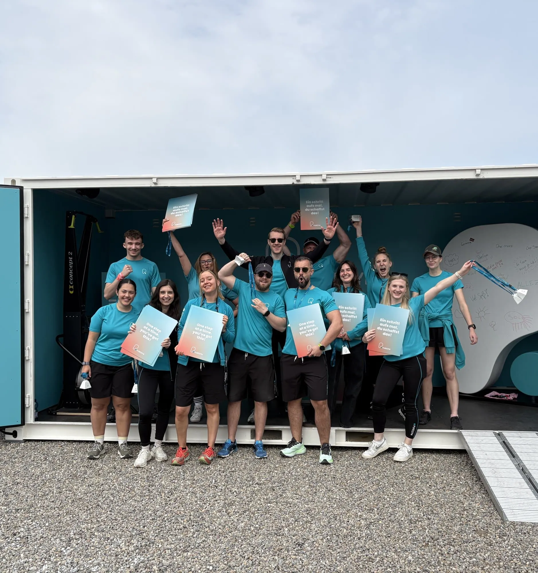 Team in türkisen PureGym-Shirts posiert mit Schildern «Ein Schritt aufs Mal, du schaffst das!» vor dem offenen Container