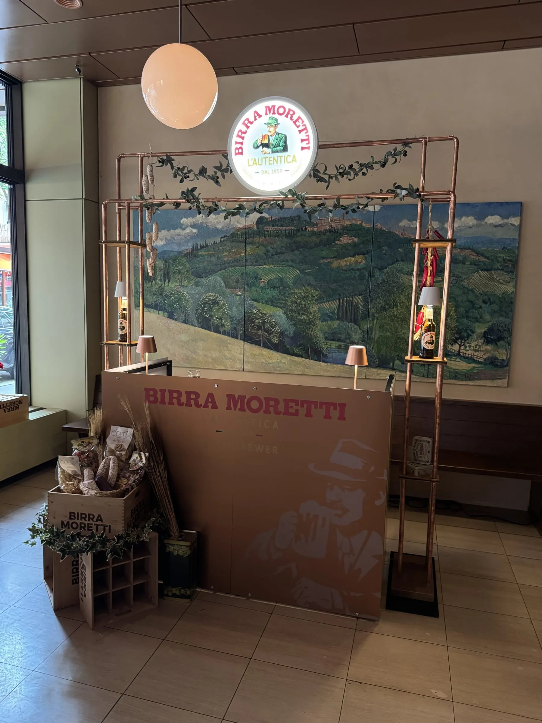 Frontansicht der Theke mit Kupferrahmen, Birra-Moretti-Leuchtschild, Salami und Peperoncini am Gestell und Holzkisten am Boden