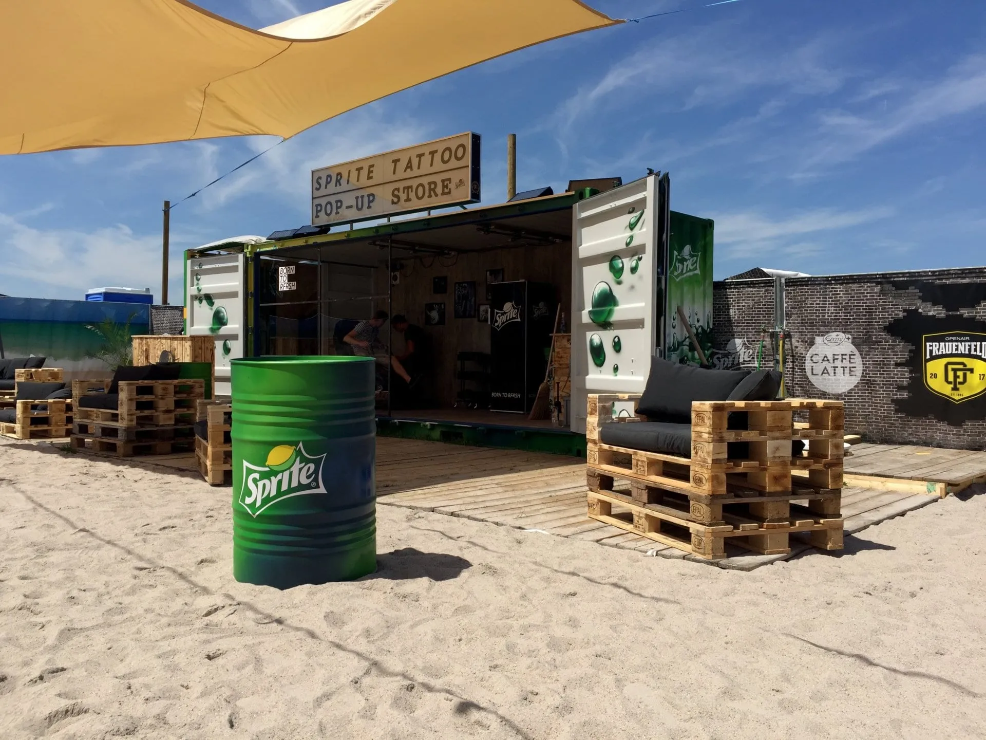 White Sprite container with an illuminated «Sprite Tattoo Pop-Up Store» sign, pallet furniture and a green barrel on sand