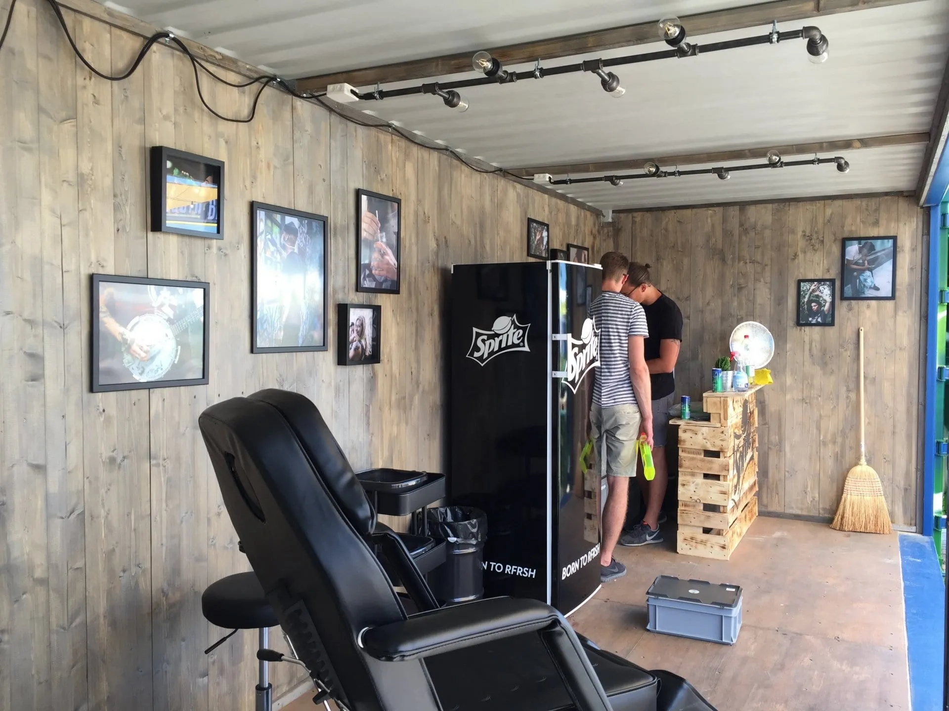 Containerinnenraum mit Holzwänden, schwarzem Tattoo-Stuhl, Sprite-Kühlschrank und gerahmten Fotos