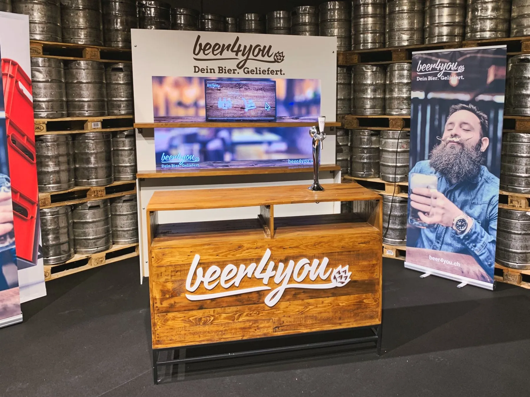 Wooden bar with beer4you lettering, a beer tap and a screen in front of stacked beer barrels