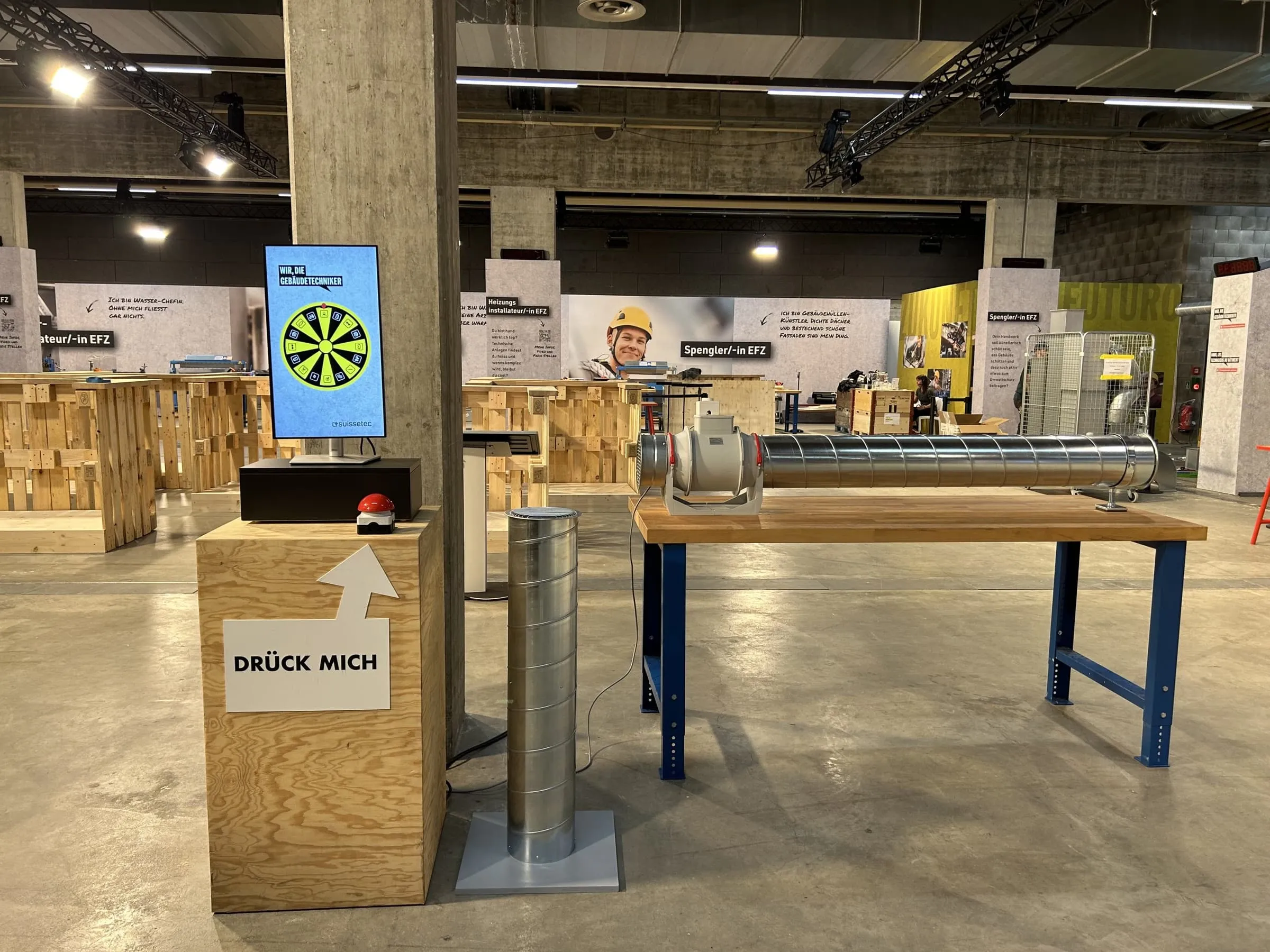 Digitales Glücksrad auf einem Screen, roter Buzzer mit Schild «Drück mich», daneben ein Lüftungsrohr auf einer Werkbank