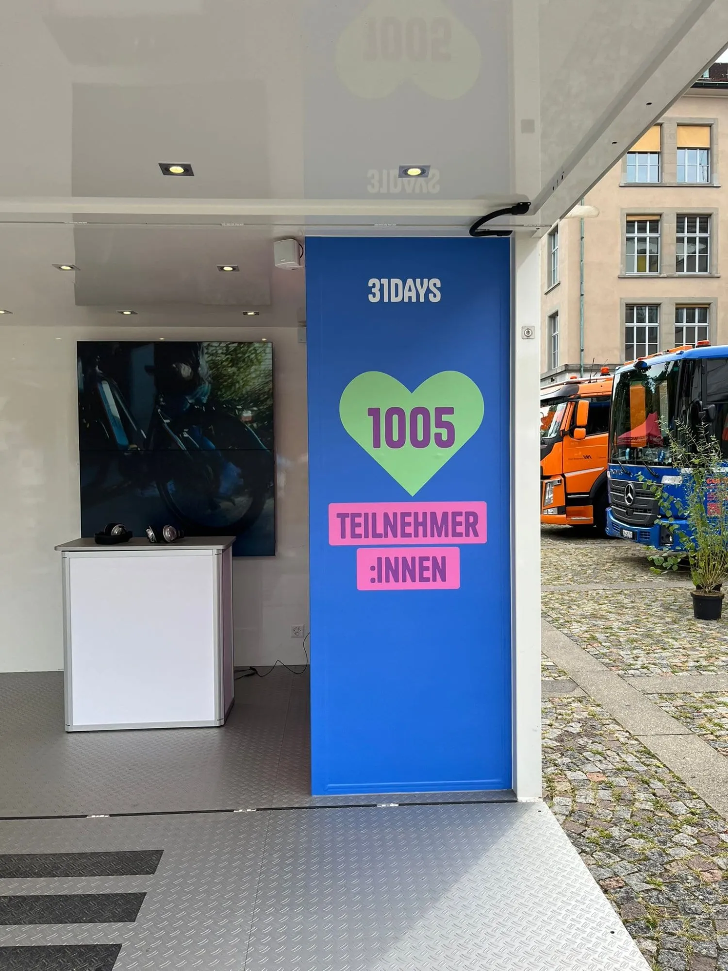 Innenraum des Cubes mit blauer 31DAYS-Wand, Theke mit Kopfhörern und Bildschirm, daneben Lastwagen