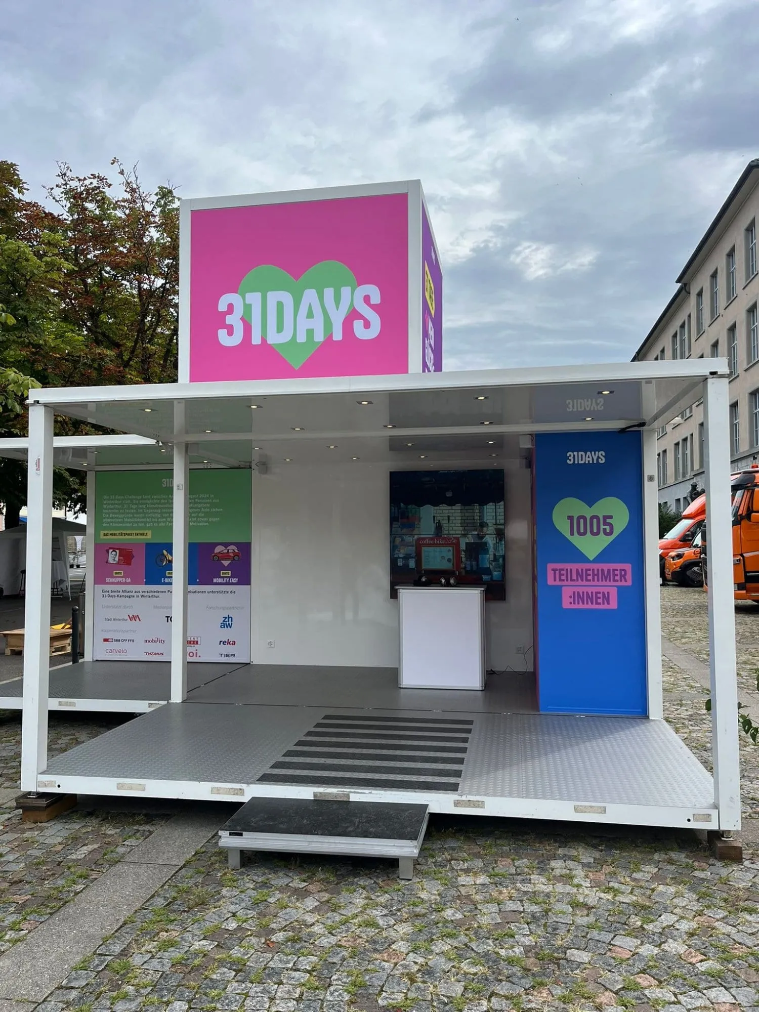 Offener Promocube mit 31DAYS-Würfel, Theke, Bildschirm und Sponsorentafel bei bewölktem Himmel