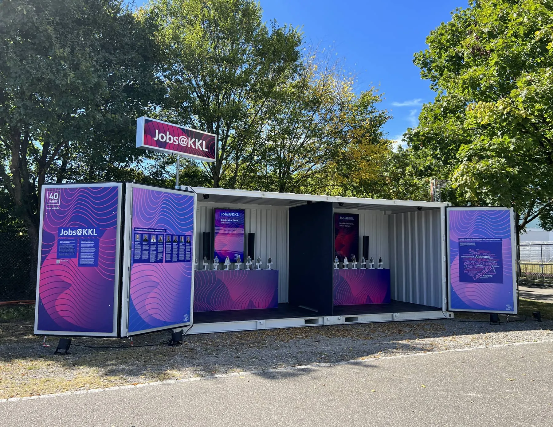 Jobs@KKL-Container mit offenen, violett-pink bedruckten Türen und zwei Bildschirmen bei Tag