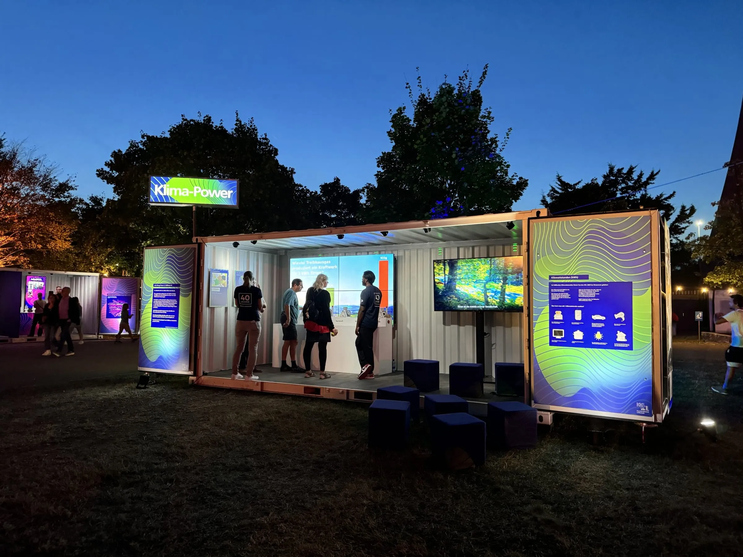 Beleuchteter Klima-Power-Container am Abend mit Besuchern vor Videowand und blauen Sitzwürfeln