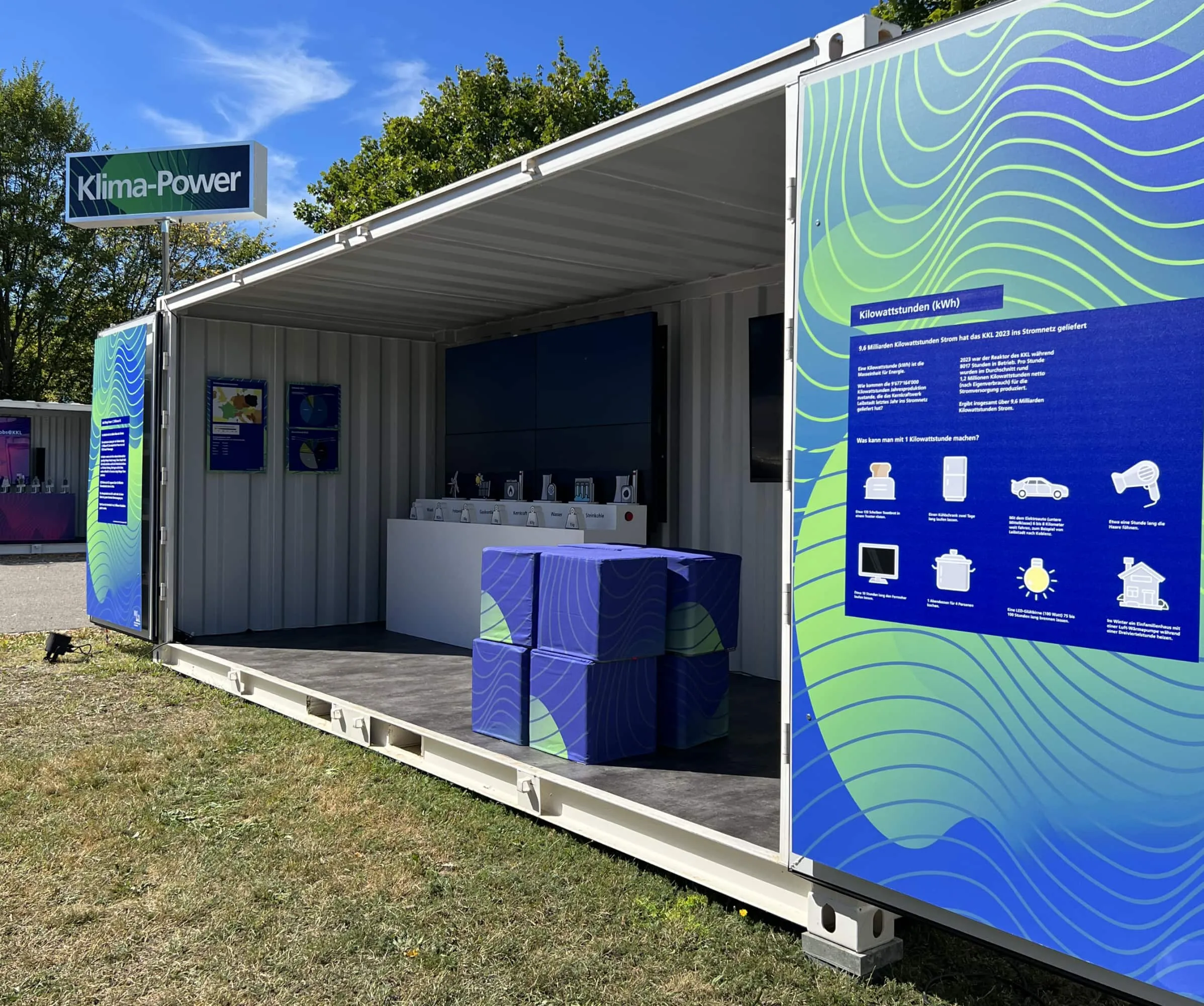 Klima-Power-Container bei Tag mit blauen Sitzwürfeln und Infotafel zu Kilowattstunden auf der Türe