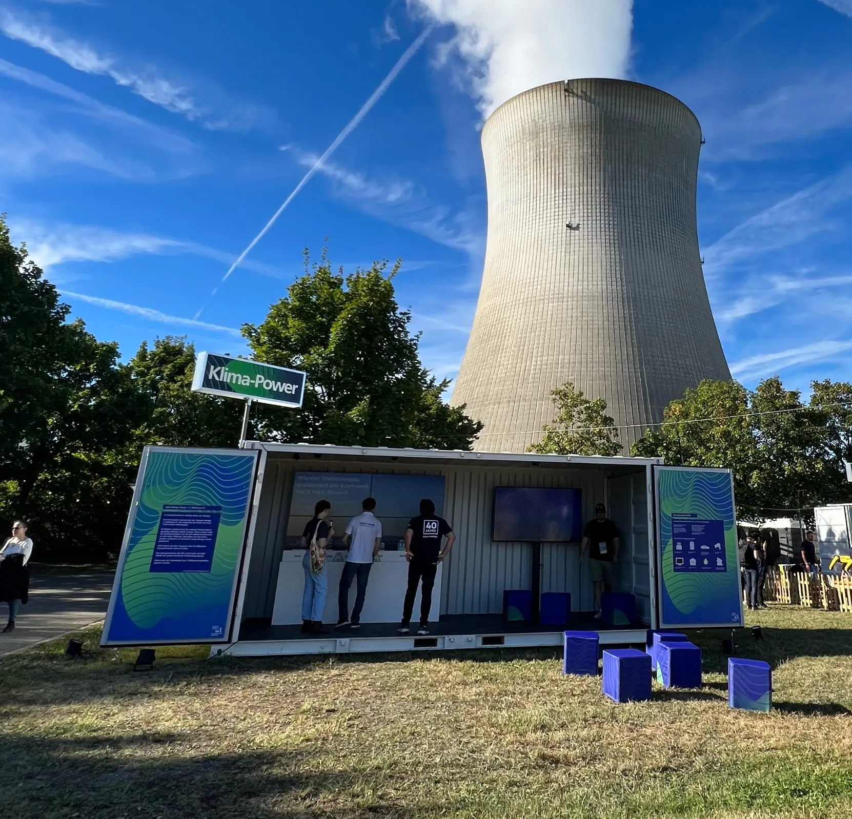 Klima-Power-Container mit offenen Türen vor dem dampfenden Kühlturm, Besucher vor der Videowand