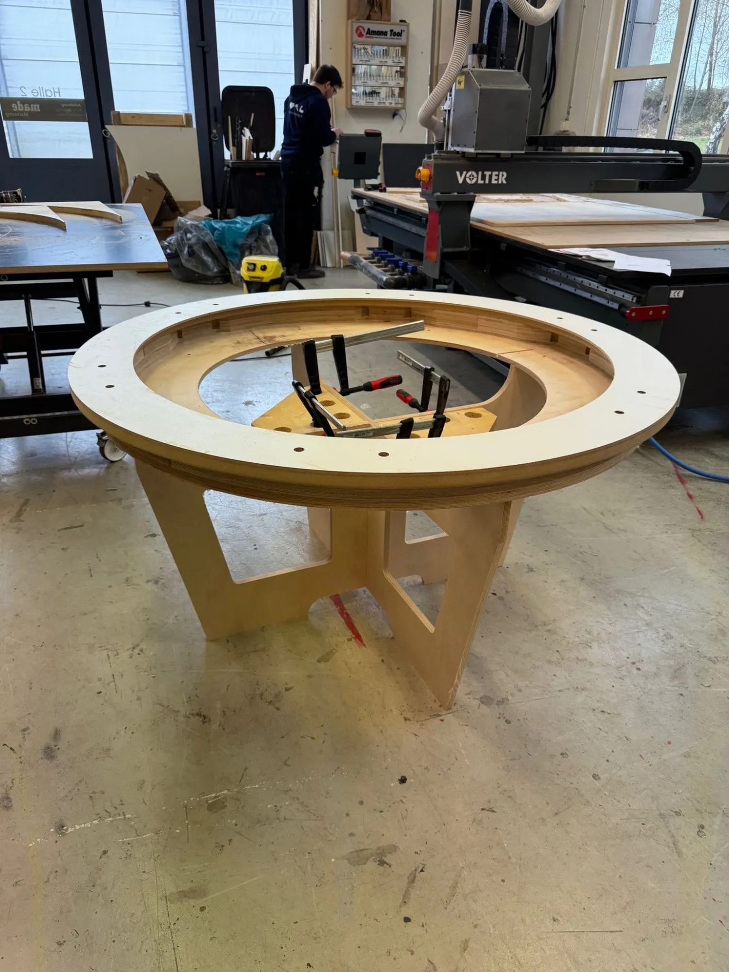 Rundes Holzgestell des Spieltischs mit Schraubzwingen in der Werkstatt, dahinter eine CNC-Fräse