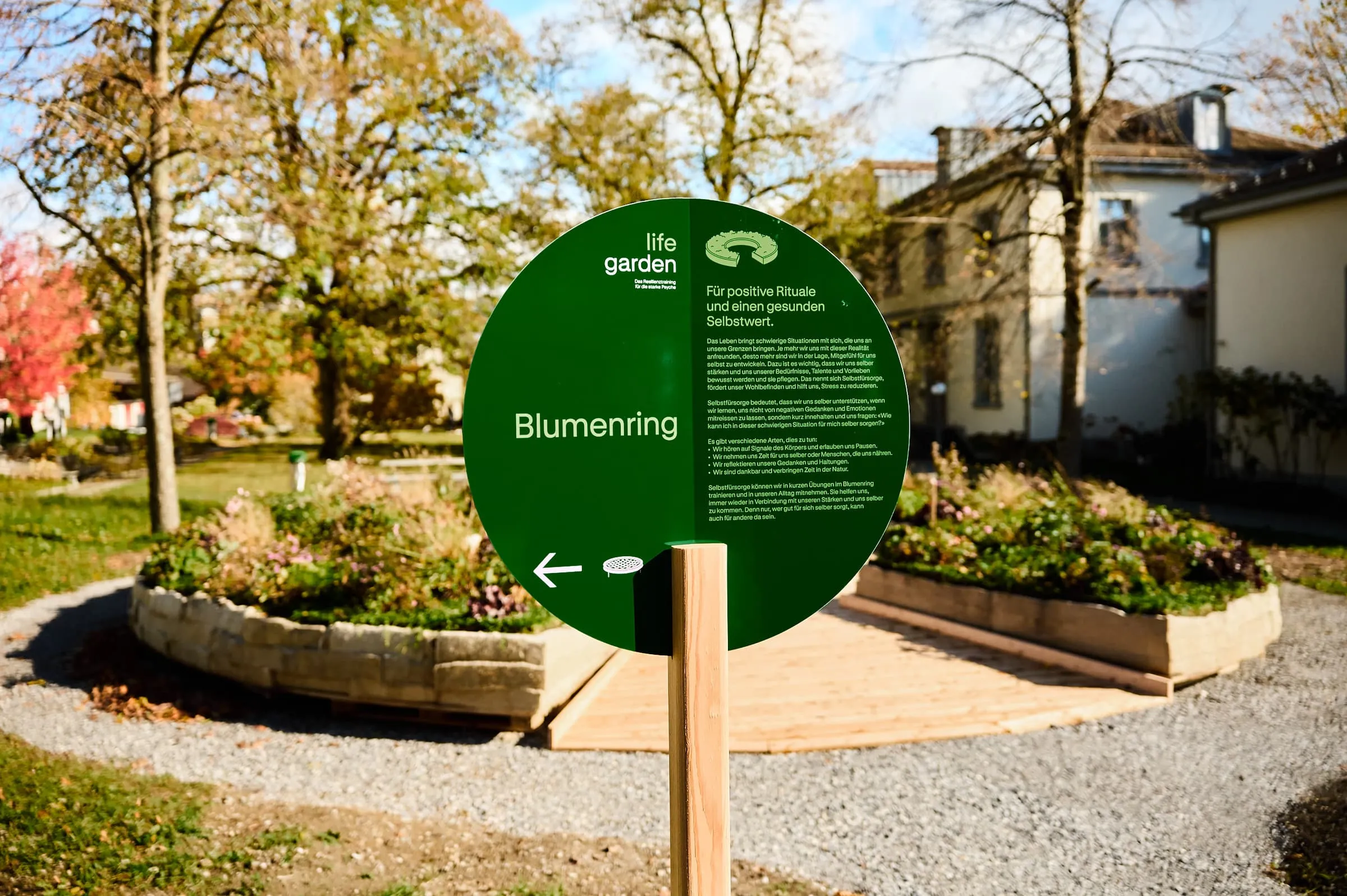 Rundes grünes Lifegarden-Schild «Blumenring» auf Holzpfosten vor dem bepflanzten Ring