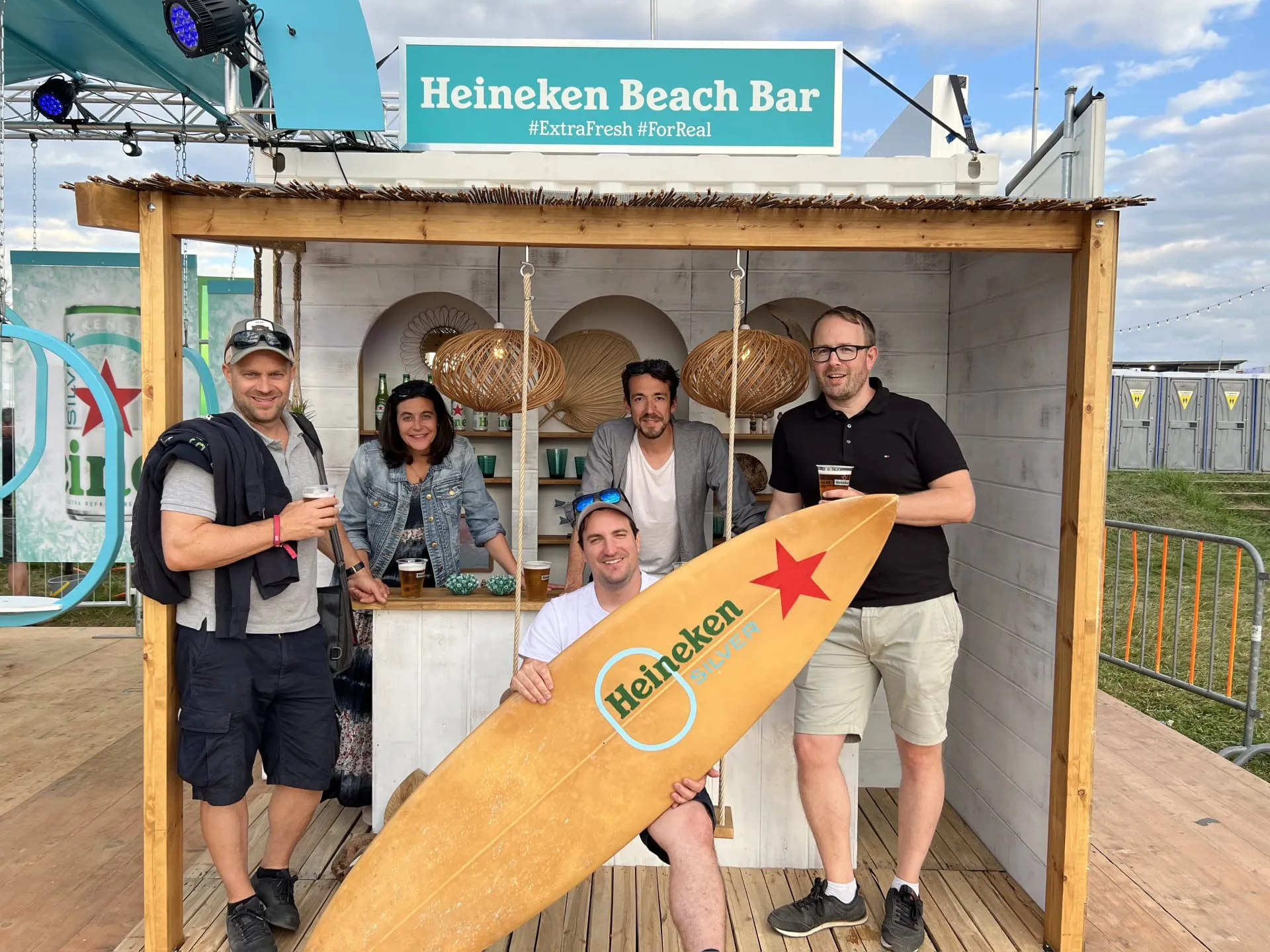 Fünf Personen mit Bier und Heineken-Surfbrett vor der Heineken Beach Bar unter dem Strohdach