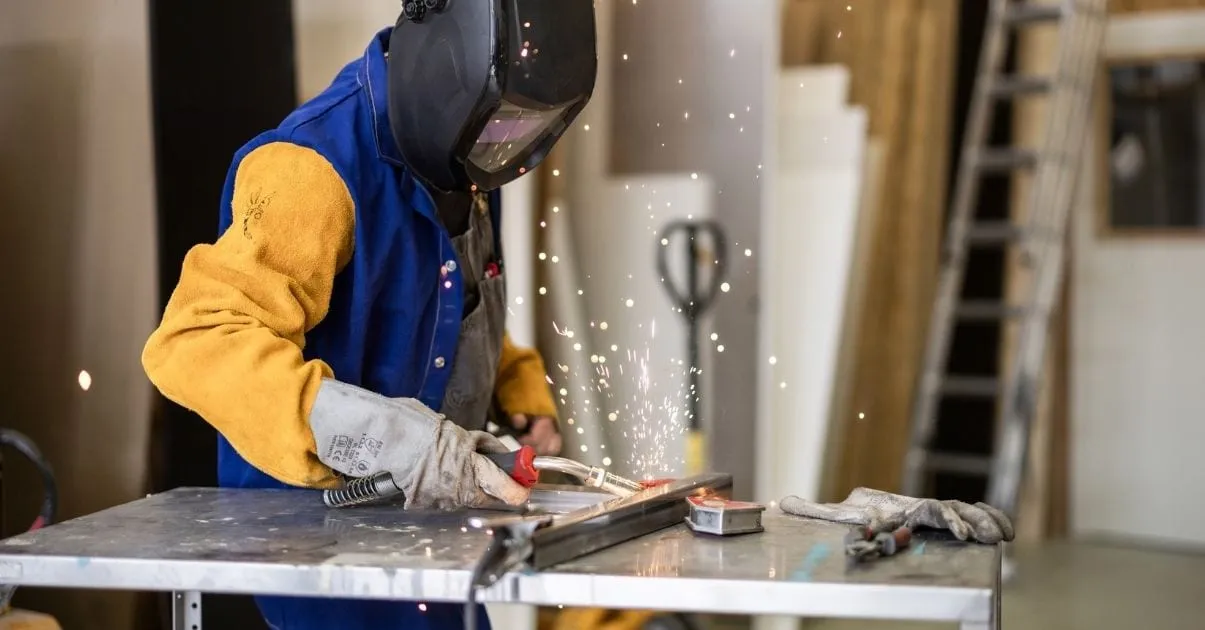 Person in a welding helmet and yellow leather sleeves welding at a metal table, sparks flying