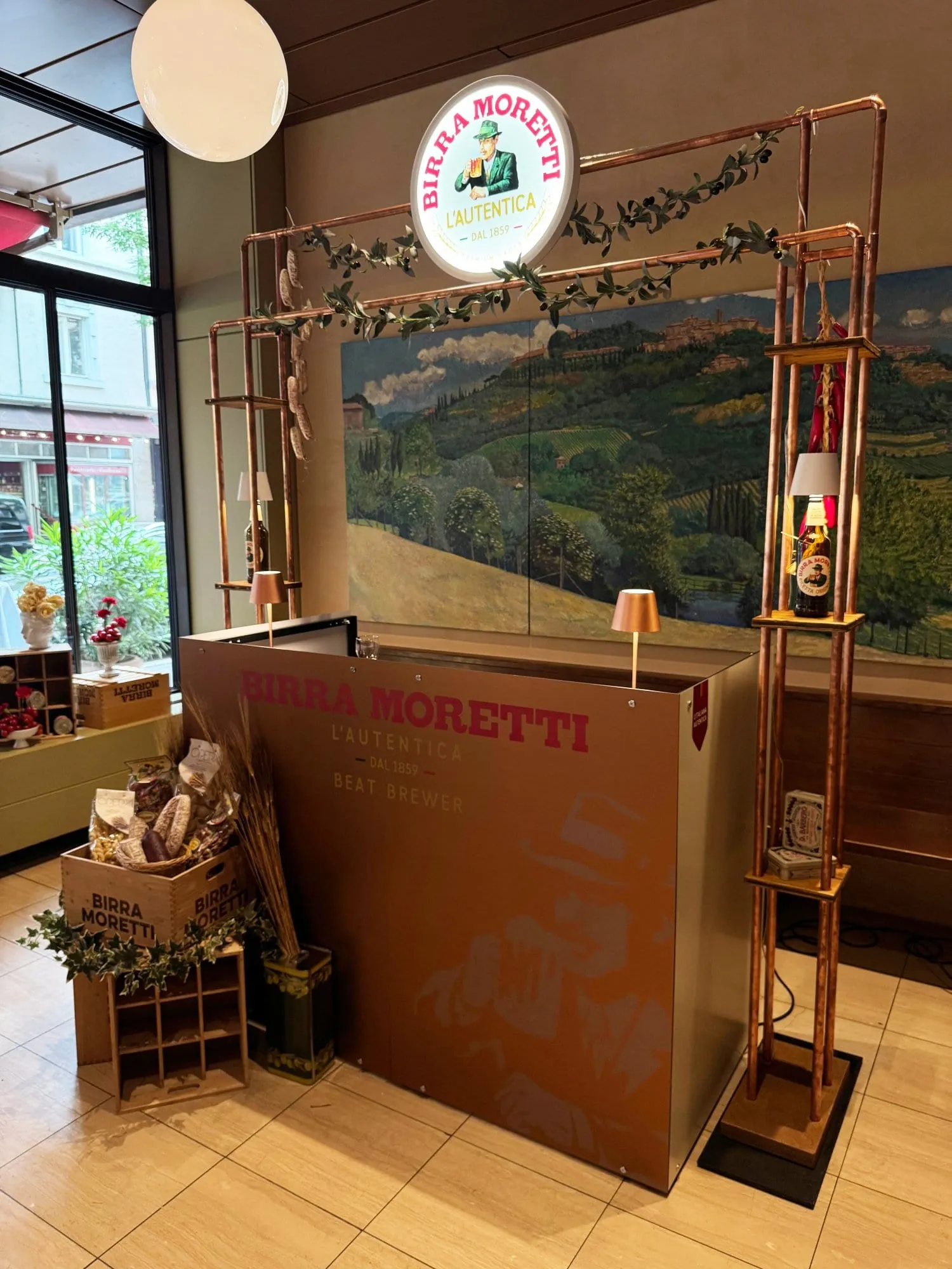 Beat-Brewer-Theke mit Kupfergestell, leuchtendem Birra-Moretti-Logo und Olivenzweigen vor einem Toskana-Wandbild im Restaurant