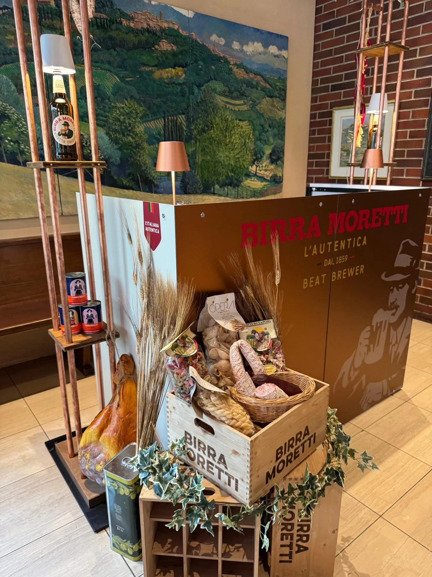 Side view of the Birra Moretti counter with a copper frame, beer-bottle lamp, wooden pasta crate and Tuscan wall image