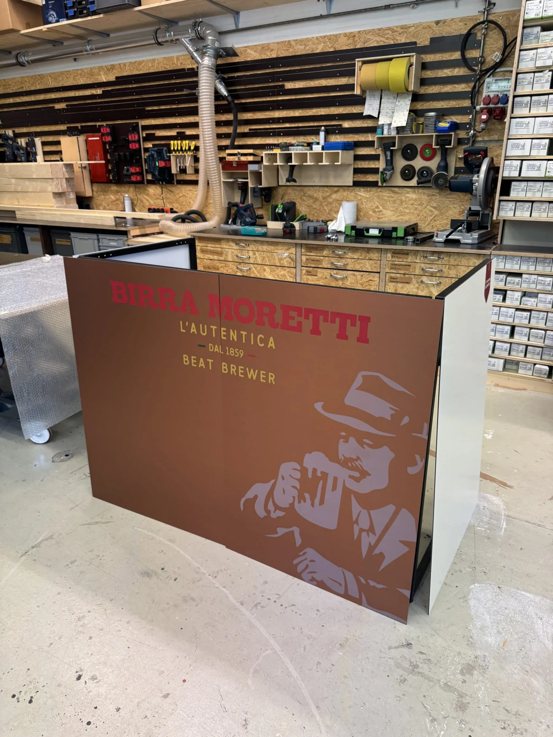 Braune Thekenverkleidung mit Birra-Moretti-Schriftzug, «Beat Brewer» und Mann-mit-Hut-Motiv in der Schreinerei