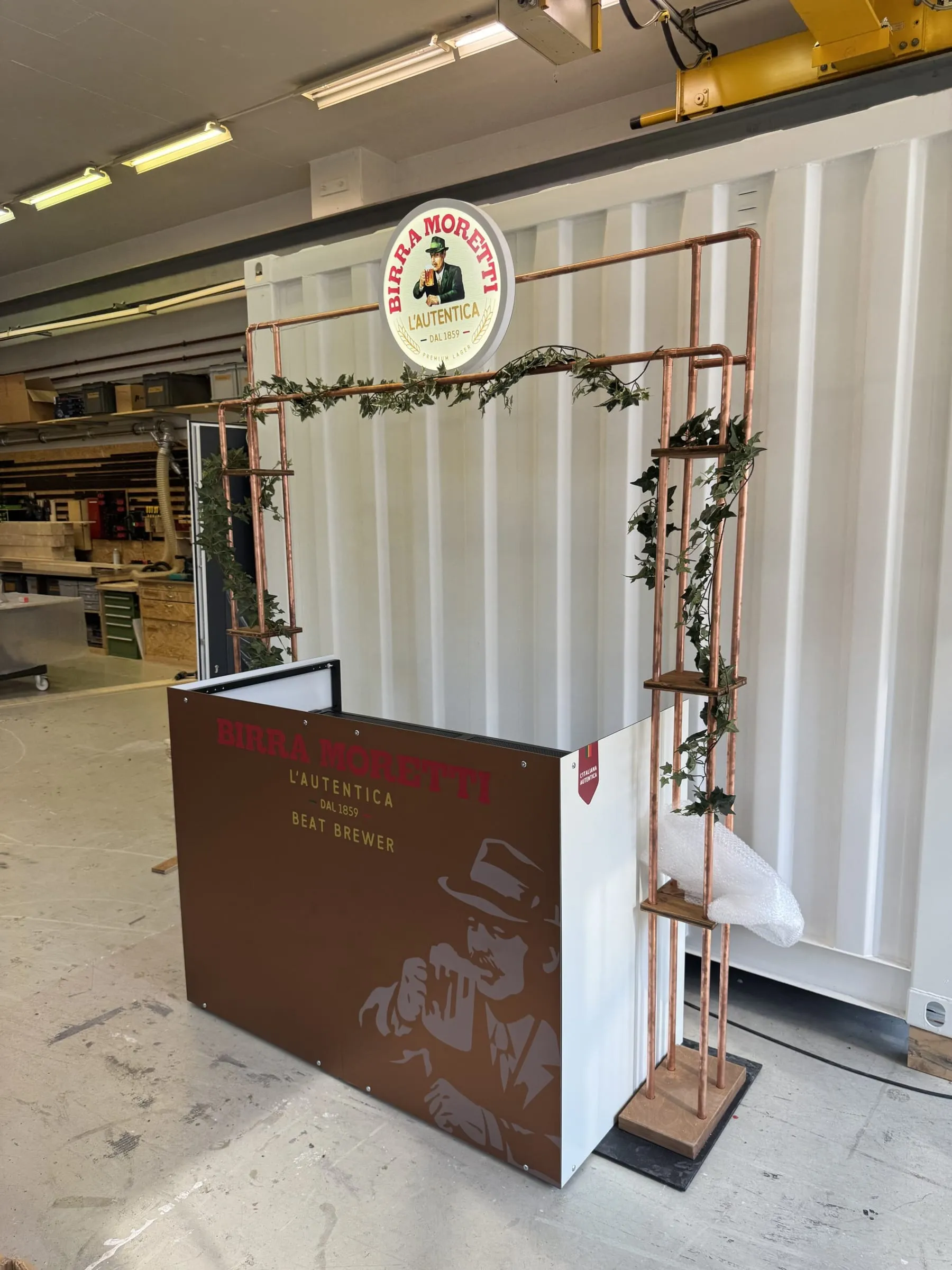 Fertige Theke mit Kupfergestell, Efeu und Leuchtschild vor einem weissen Container in der Werkhalle