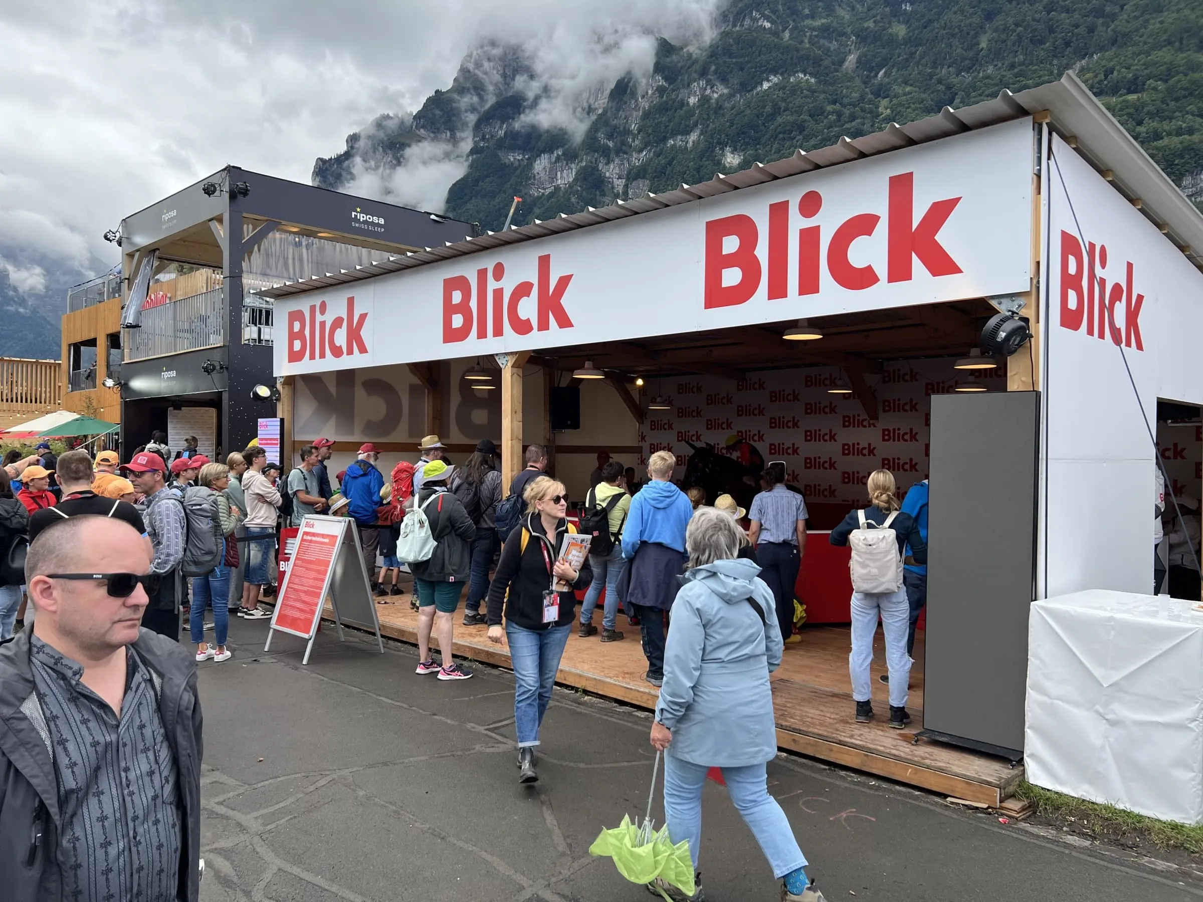 Warteschlange vor dem Blick-Unterstand mit rotem Rodeo-Ring, im Hintergrund wolkenverhangene Berge