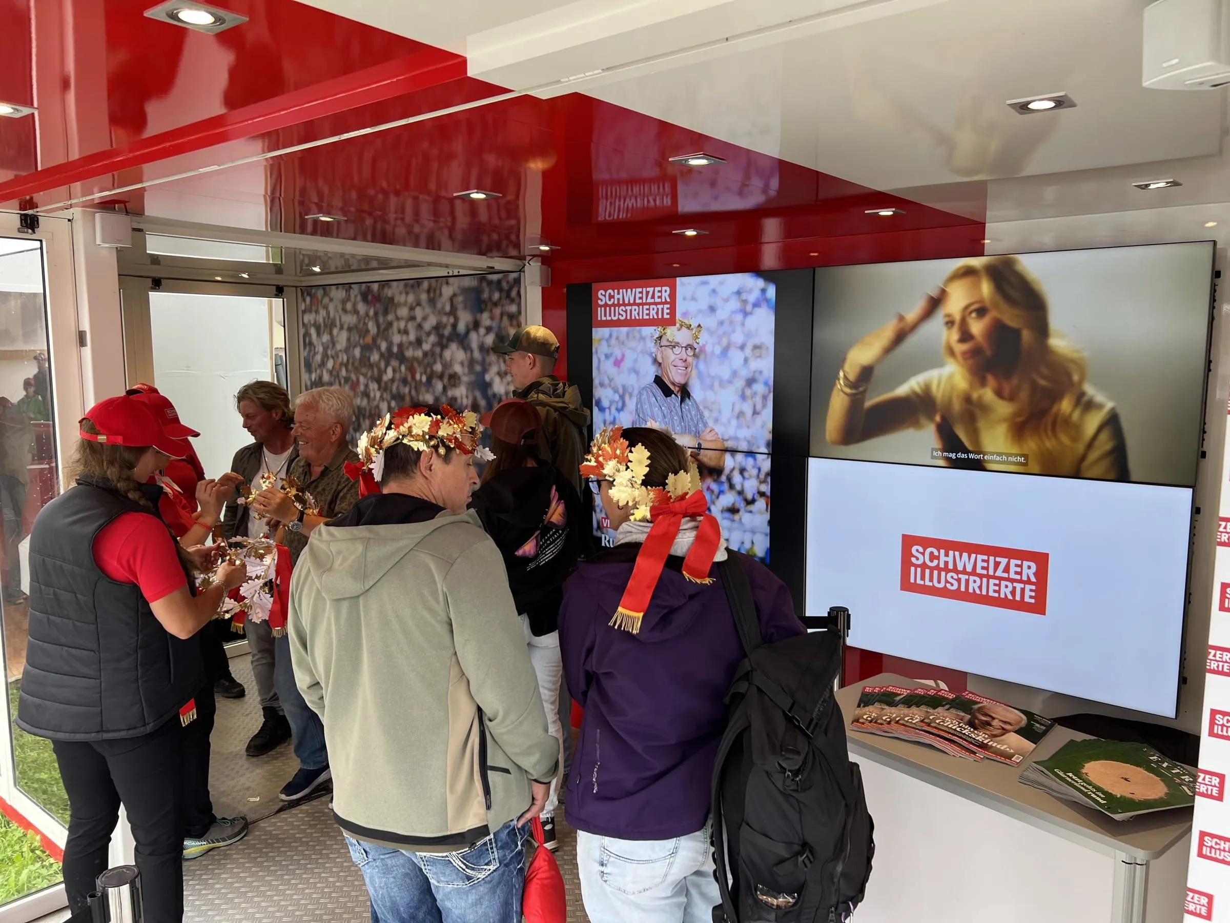 Gäste mit Eichenlaub-Kränzen im SI-Container, an der Wand Screens mit SI-Cover und Video