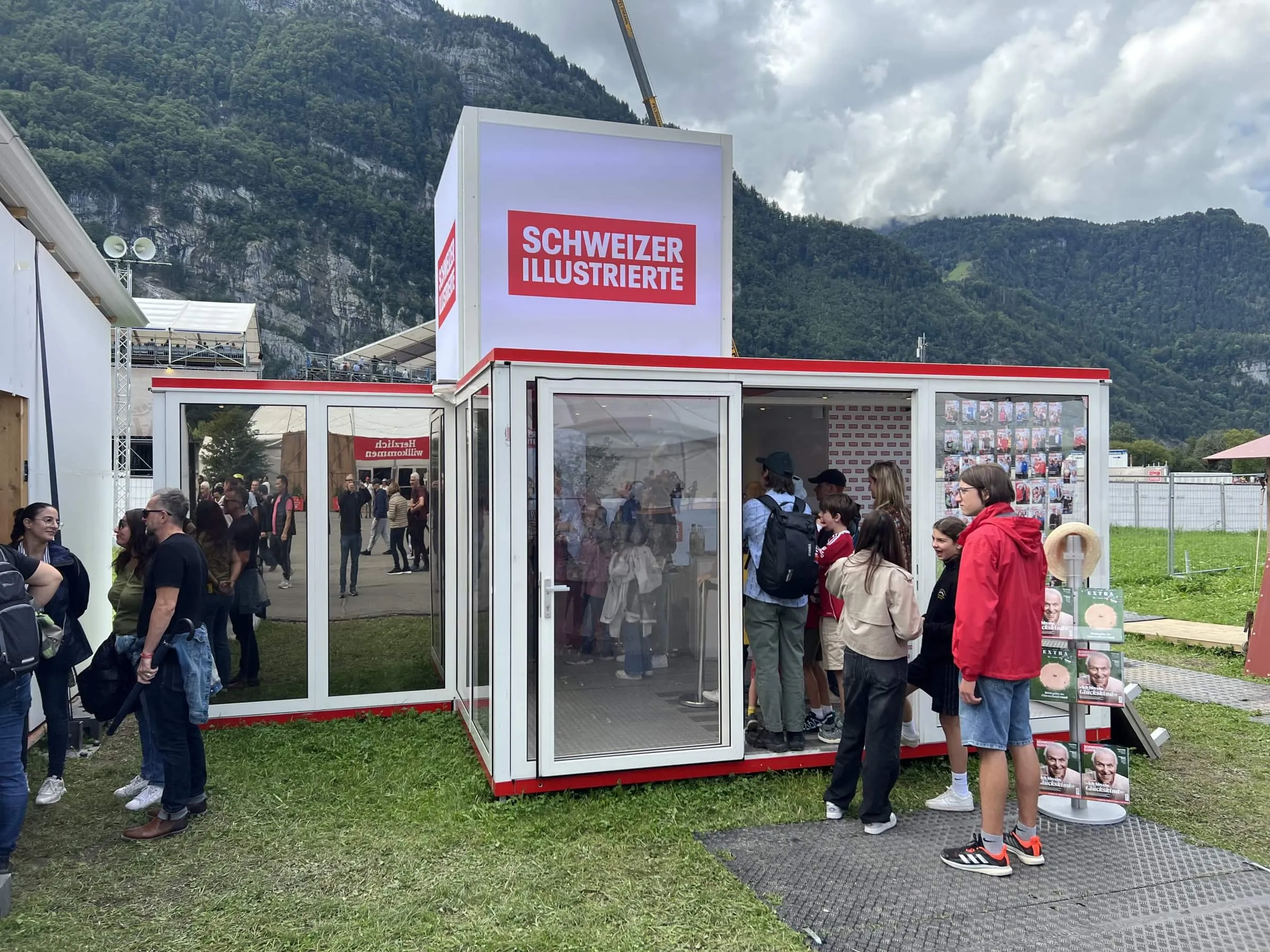 SI-Glascontainer mit Leuchtwürfel Schweizer Illustrierte, Besuchende warten am Eingang