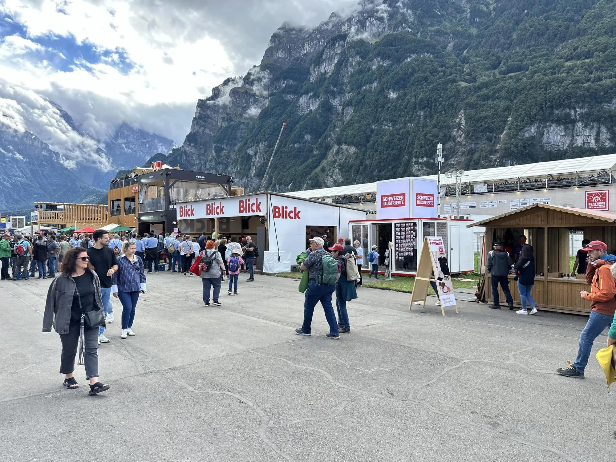 Blick-Unterstand, SI-Container und Fanshop-Hütte auf dem ESAF-Gelände in Mollis, davor Besuchende