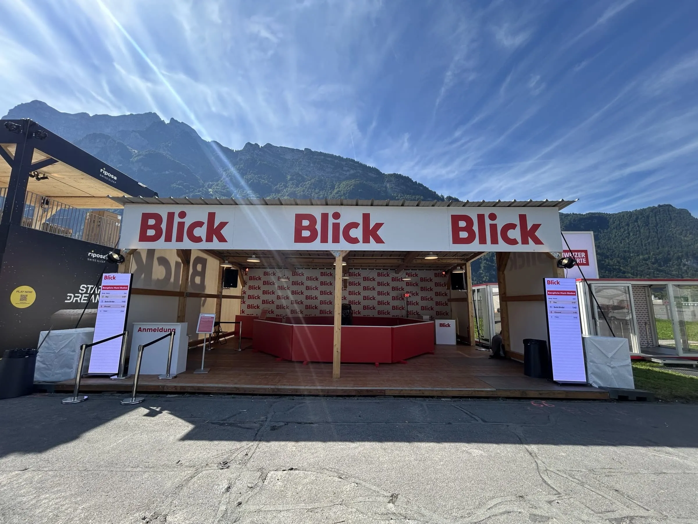 Holzunterstand mit dreifachem Blick-Schriftzug, roter Rodeo-Arena und Rangliste-Screens vor der Bergkulisse