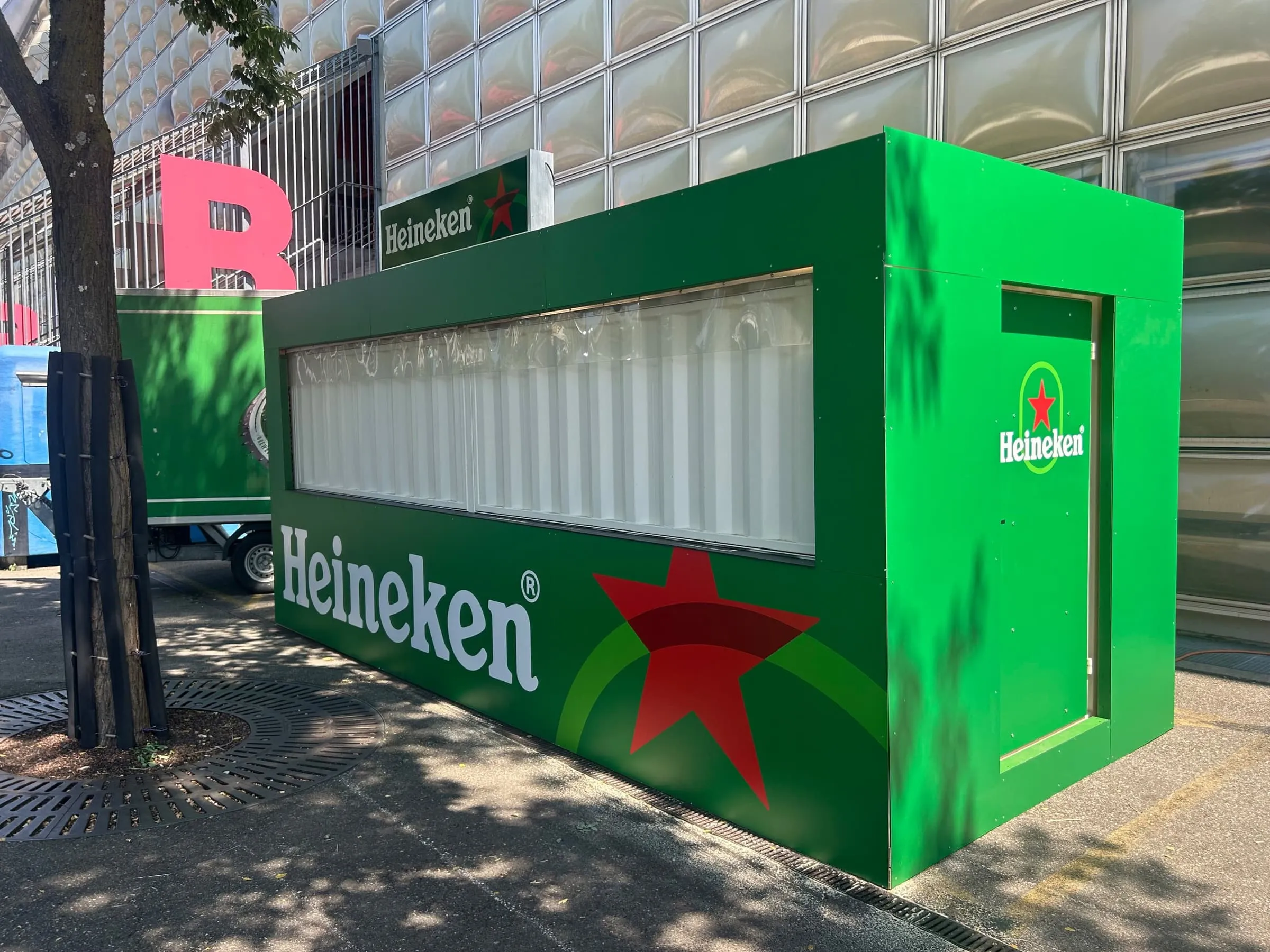 Heineken-Container mit Seitentüre und Heineken-Logo vor einer Fassade aus Glaskissen in Basel, dahinter ein grüner Anhänger