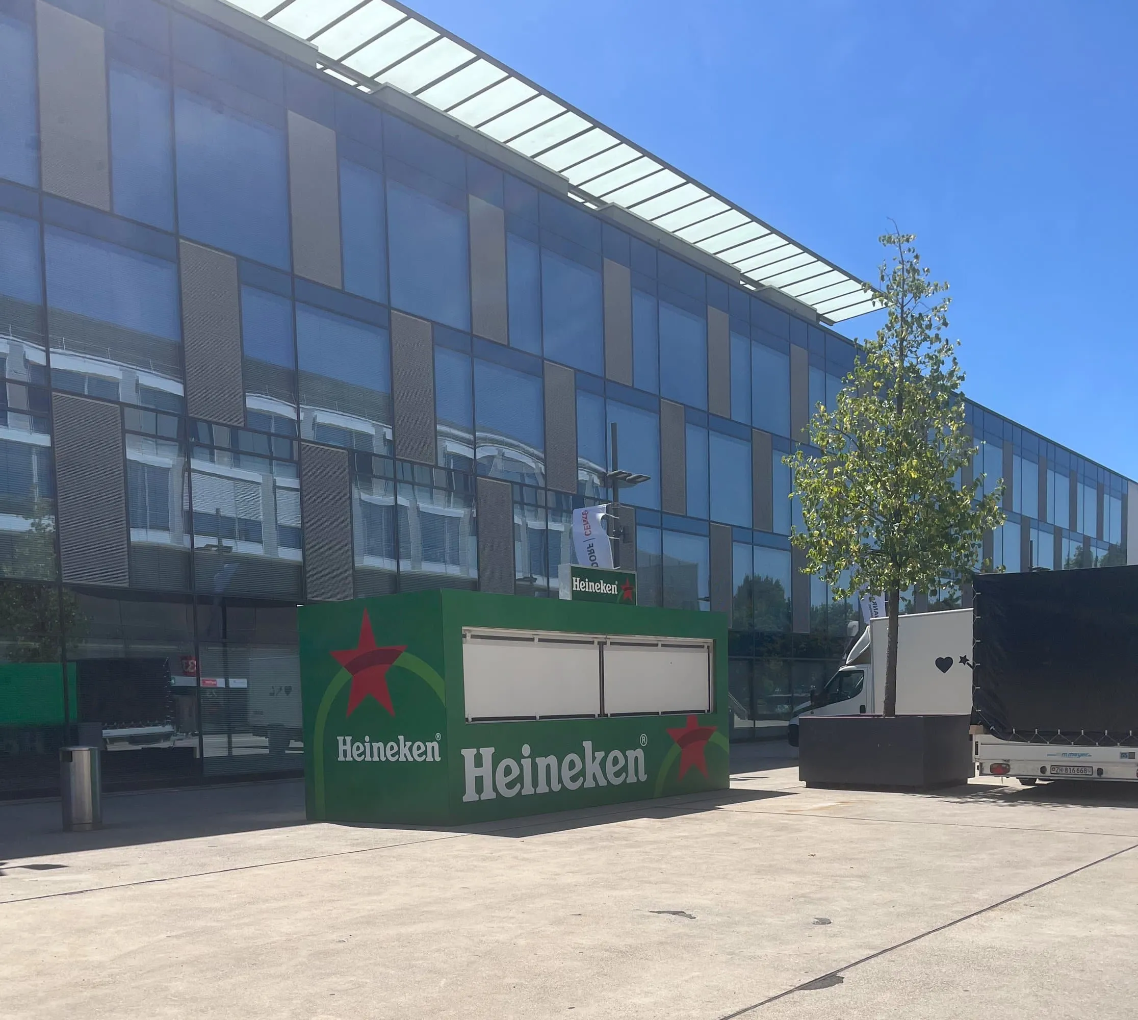 Heineken-Container vor einer Glasfassade in Bern, daneben ein Baum und ein Lieferwagen mit Anhänger