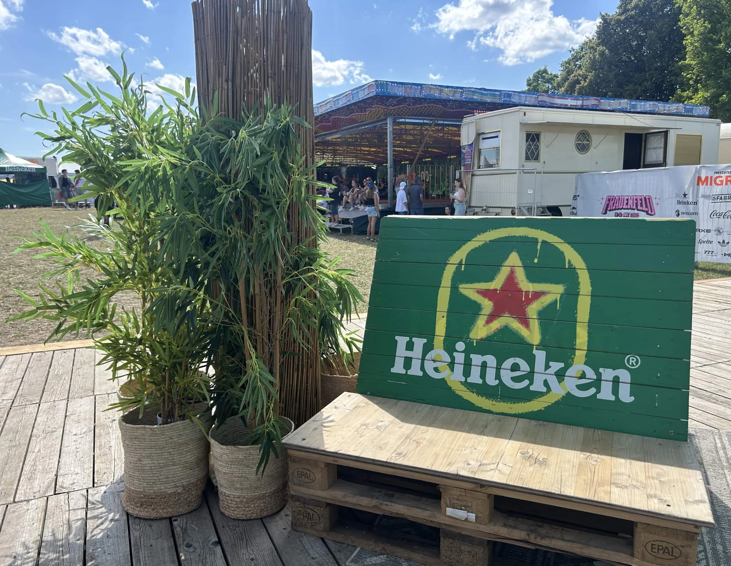 Palettenbank mit grünem Heineken-Holzschild und Bambuspflanzen, im Hintergrund Autoscooter und ein Wohnwagen