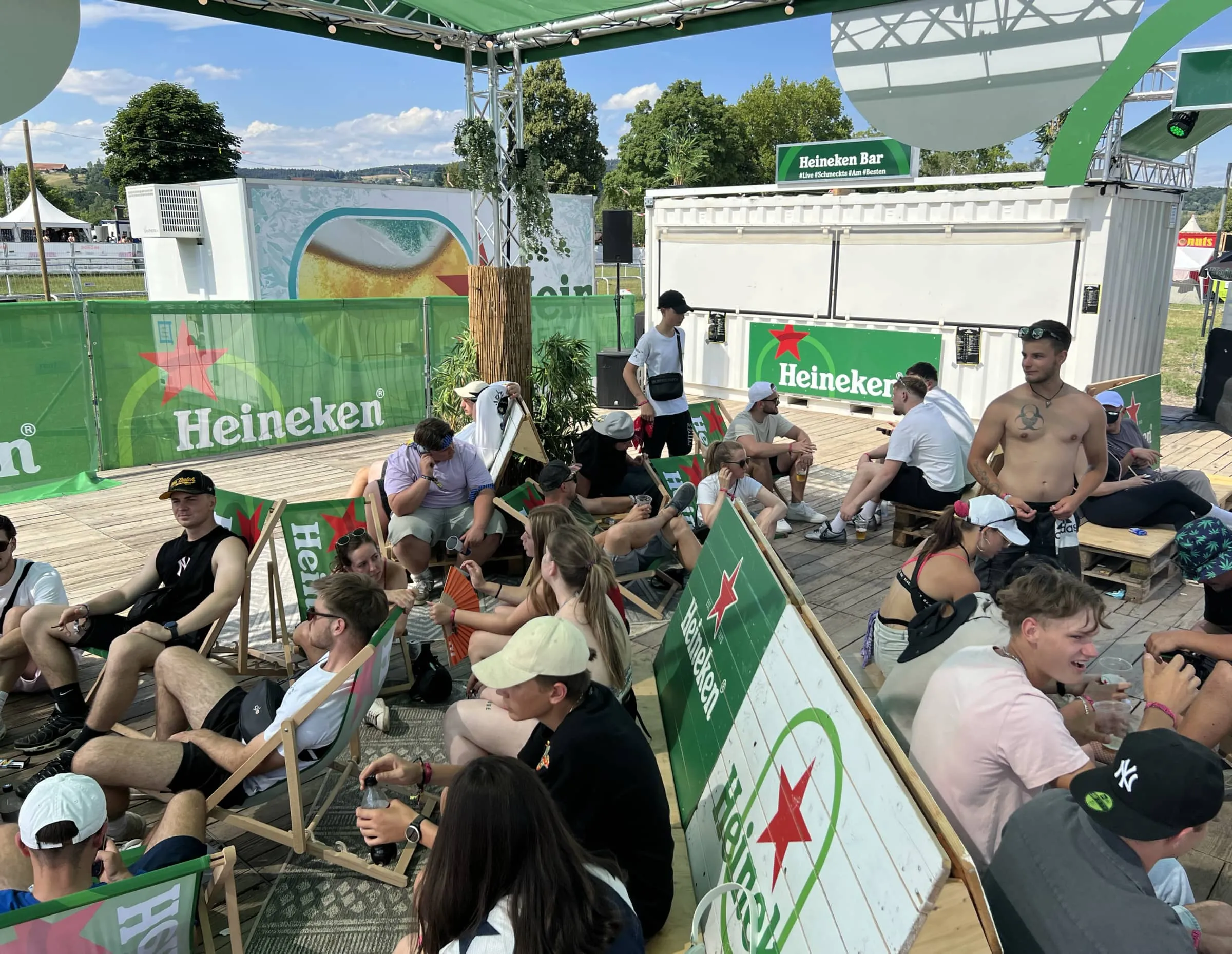 Festivalbesucher in Heineken-Liegestühlen unter dem Sonnendach, dahinter ein weisser Barcontainer mit «Heineken Bar»-Schild