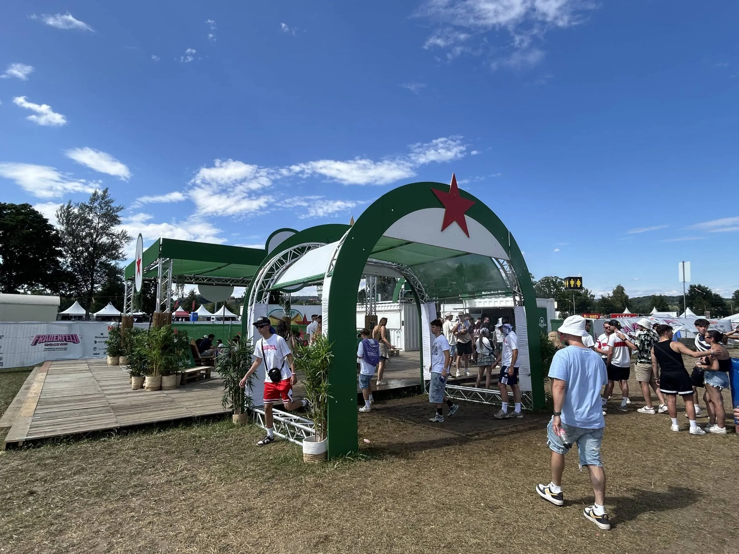Grüner Torbogen mit rotem Stern am Eingang zur Heineken-Area, Festivalbesucher auf dem Holzpodest bei Sonnenschein