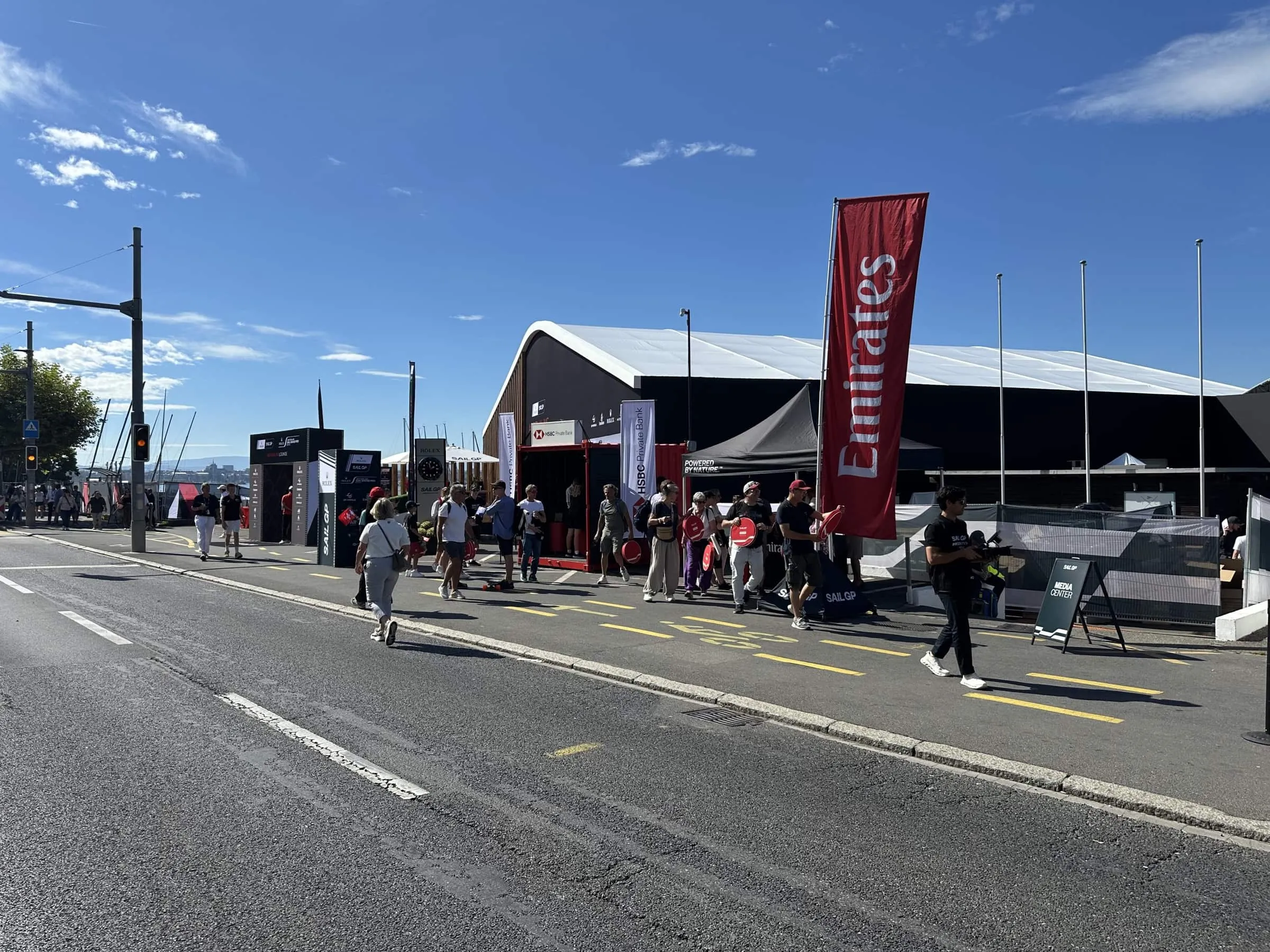 Strasse am SailGP-Gelände in Genf mit HSBC-Container, Emirates-Fahne und Besuchenden mit Fächern