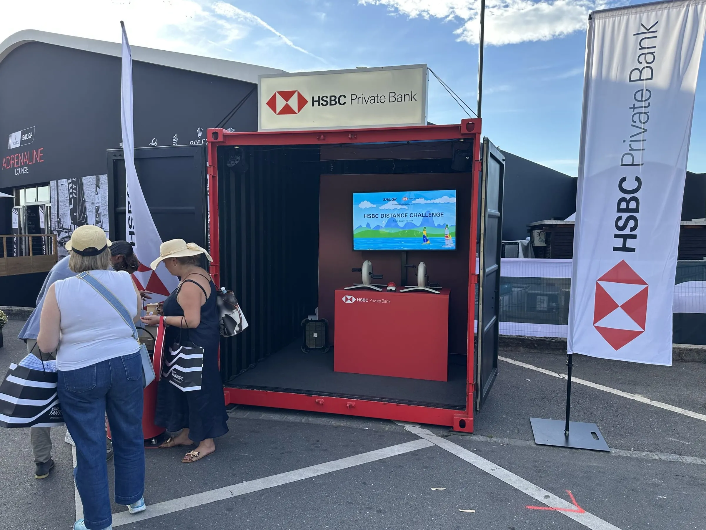Drei Besucherinnen mit Sonnenhüten vor dem HSBC-Container, der Screen zeigt die Distance Challenge