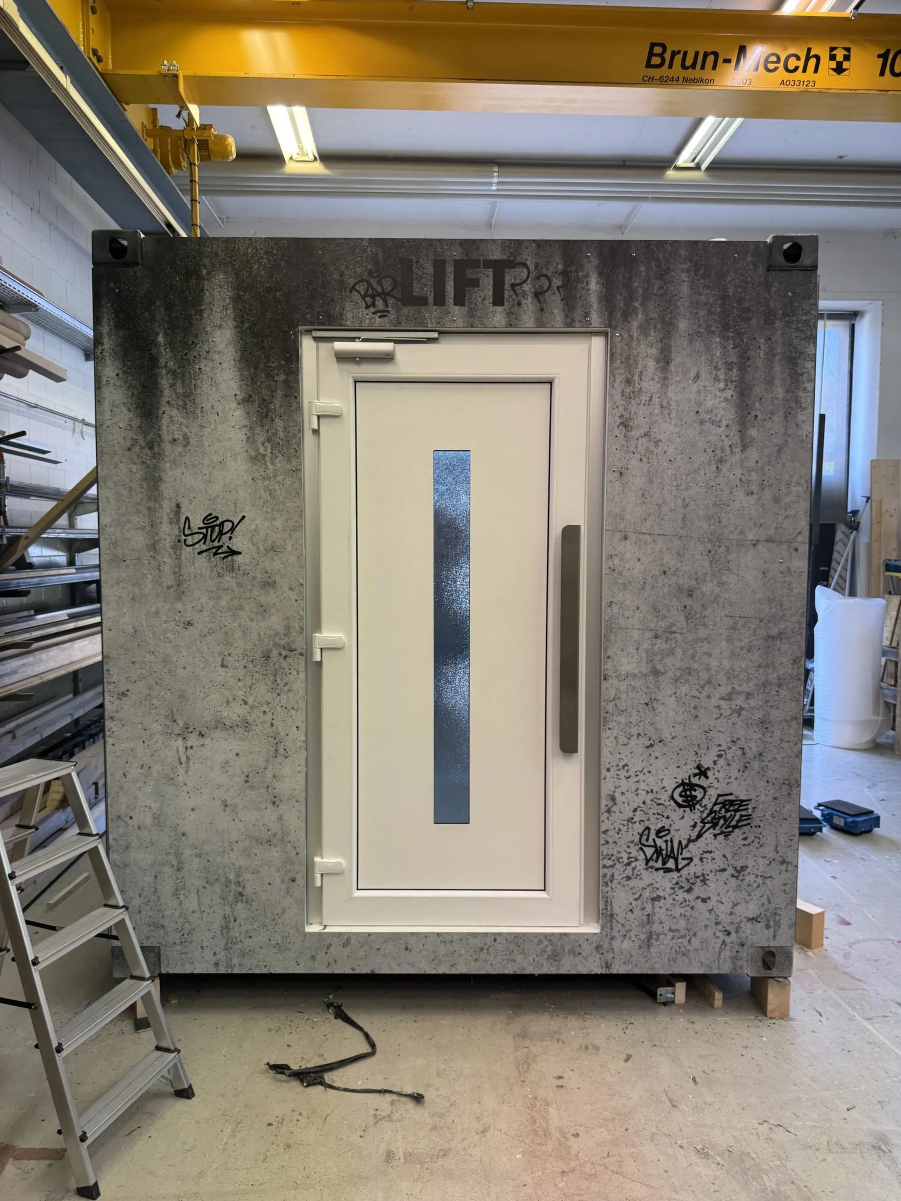 Container in der Werkstatt unter einem gelben Kranträger, Betonlook-Fassade mit weisser Lifttür und Graffiti