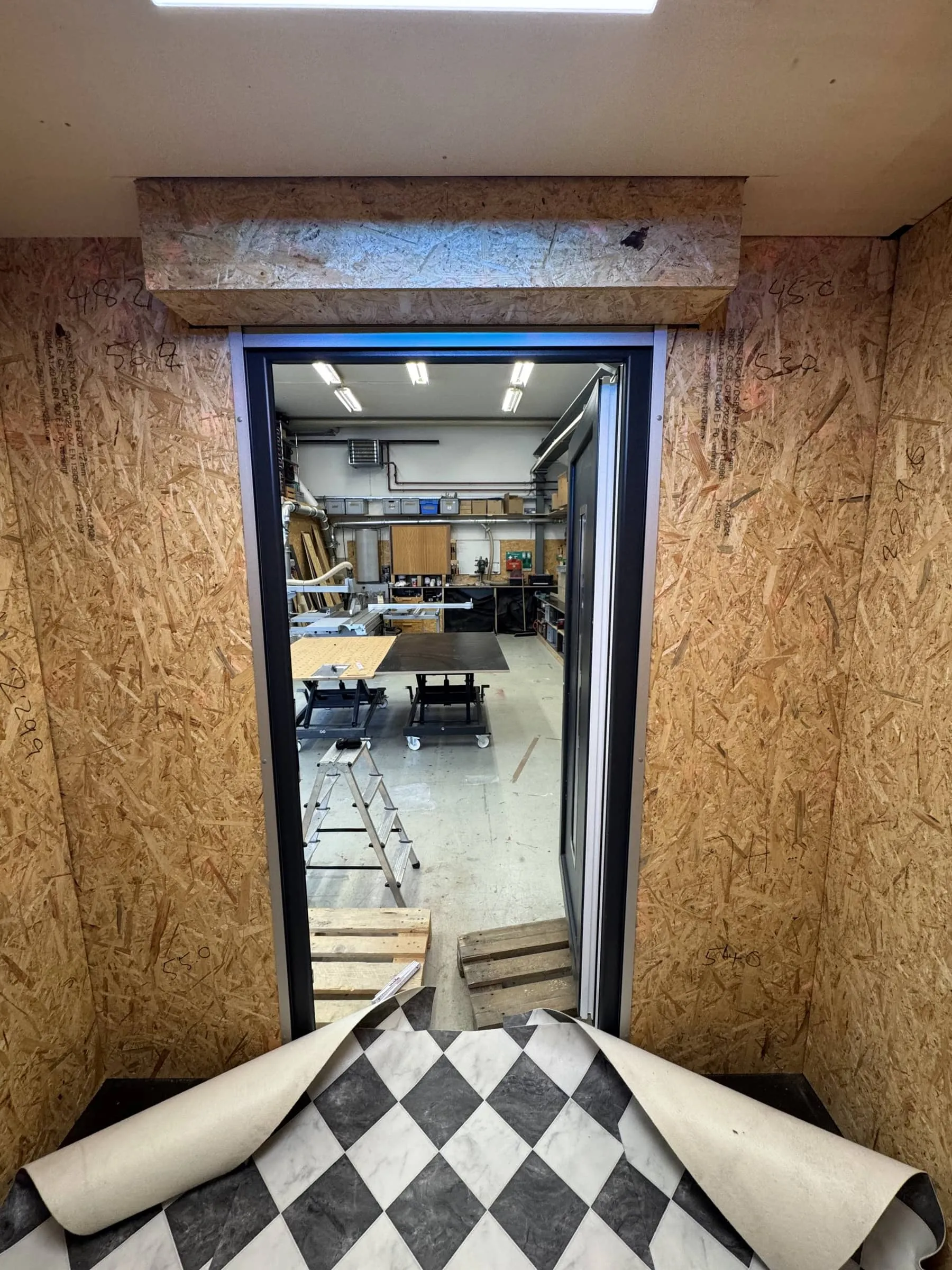 Blick aus dem OSB-verkleideten Innenraum durch die Türöffnung in die Werkstatt, Schachbrett-Bodenbelag im Vordergrund