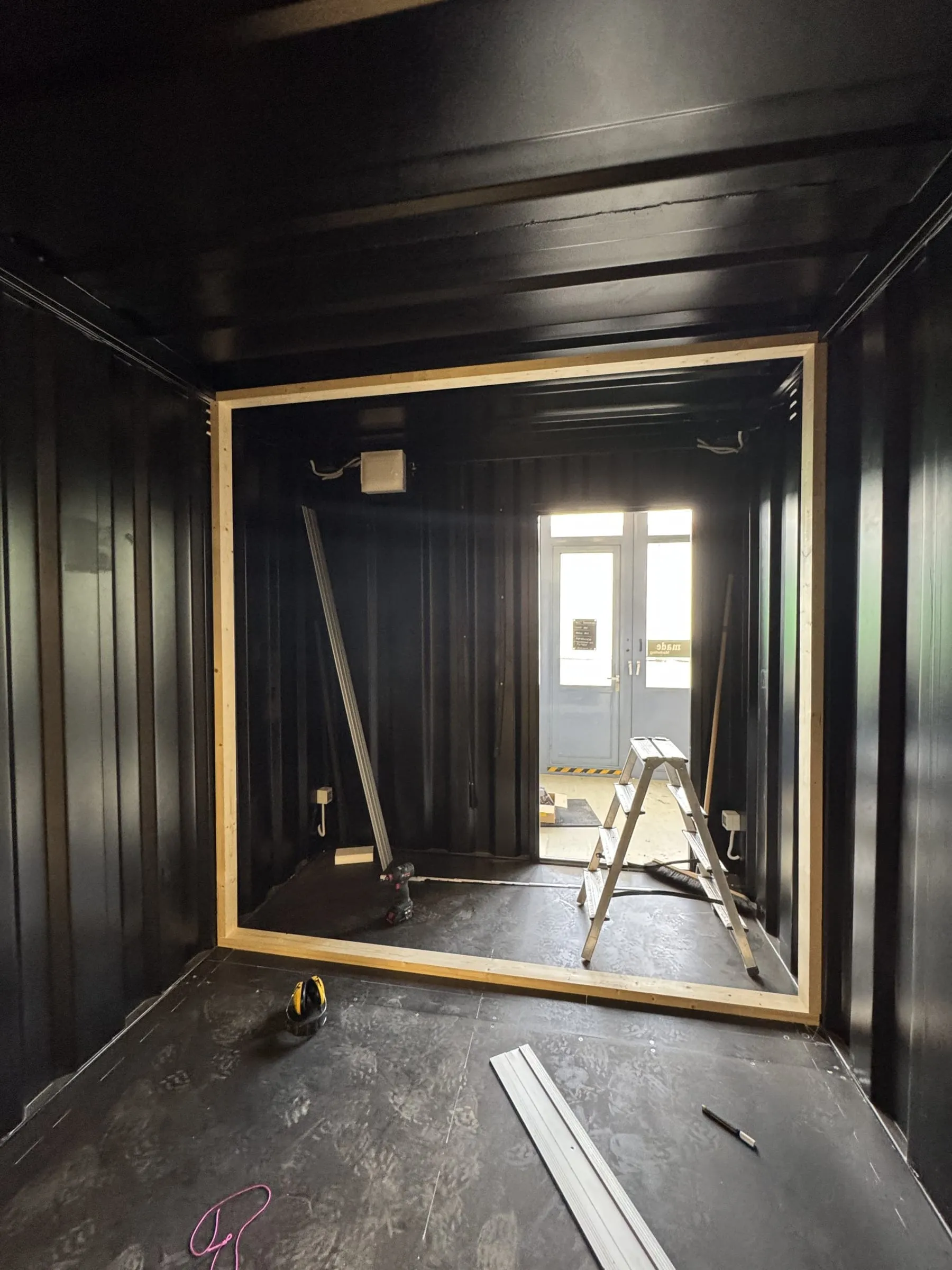 Schwarz lackierter Containerinnenraum im Rohbau mit Holzrahmen, Leiter und Akkuschrauber