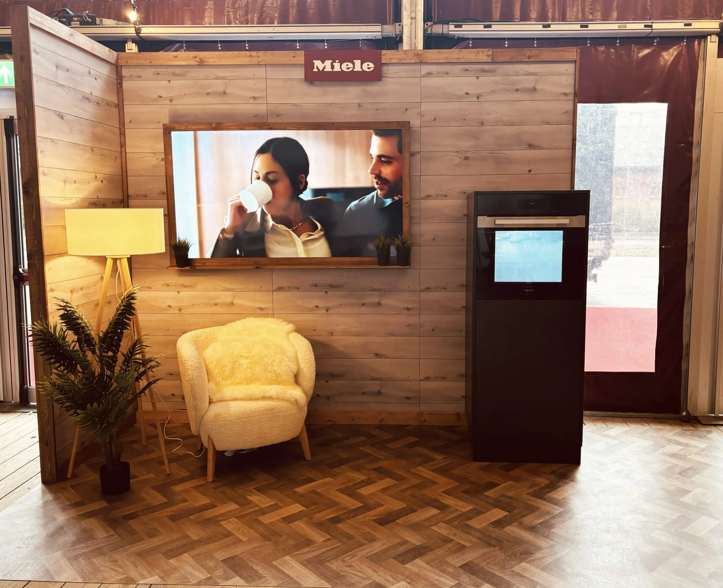 Miele stand in a wooden chalet style with a framed screen, sheepskin-covered armchair, floor lamp and oven column