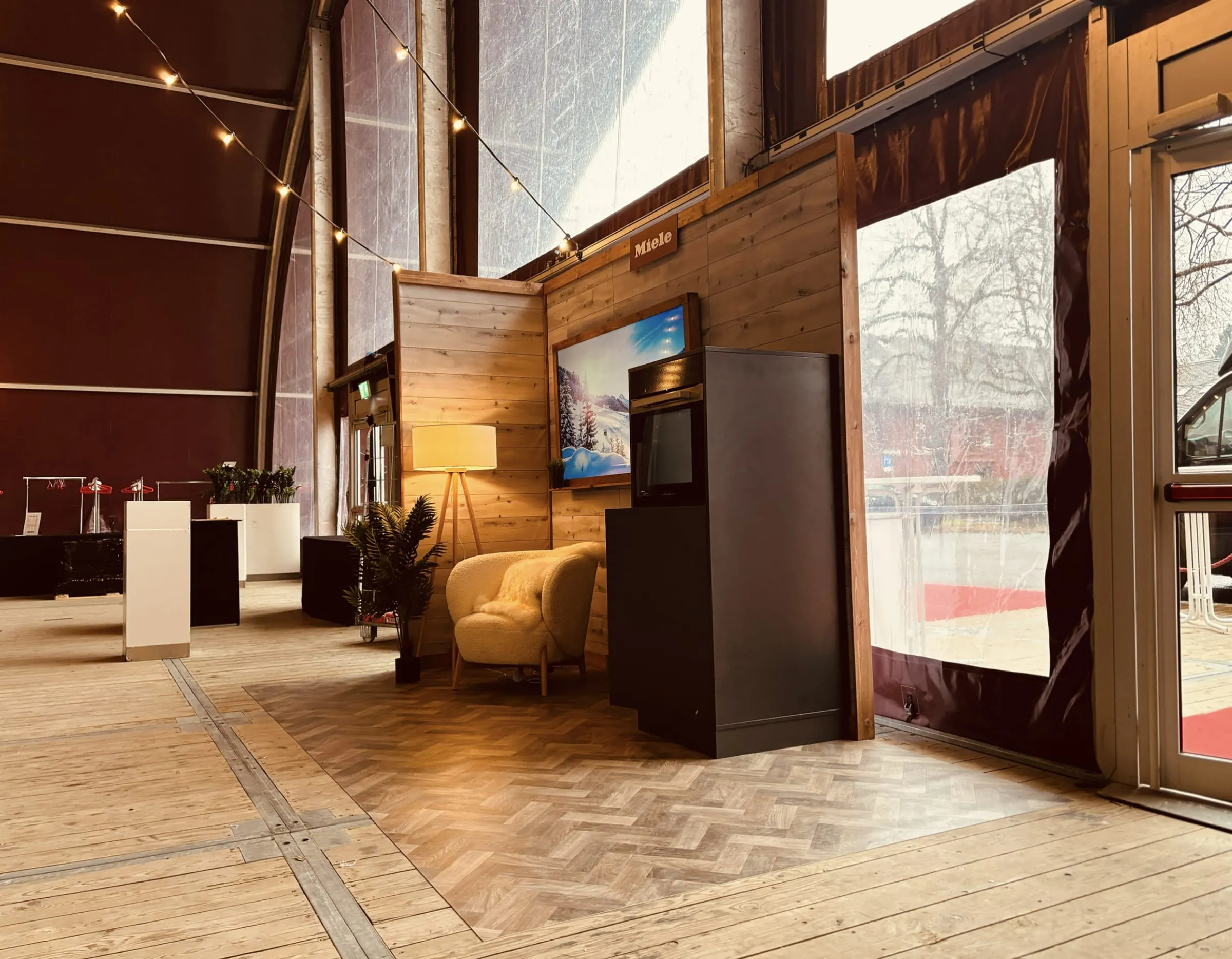 Miele-Chalet-Ecke im Zelt mit Holzwänden, Lichterketten, Winterlandschaft auf dem Screen und Backofen