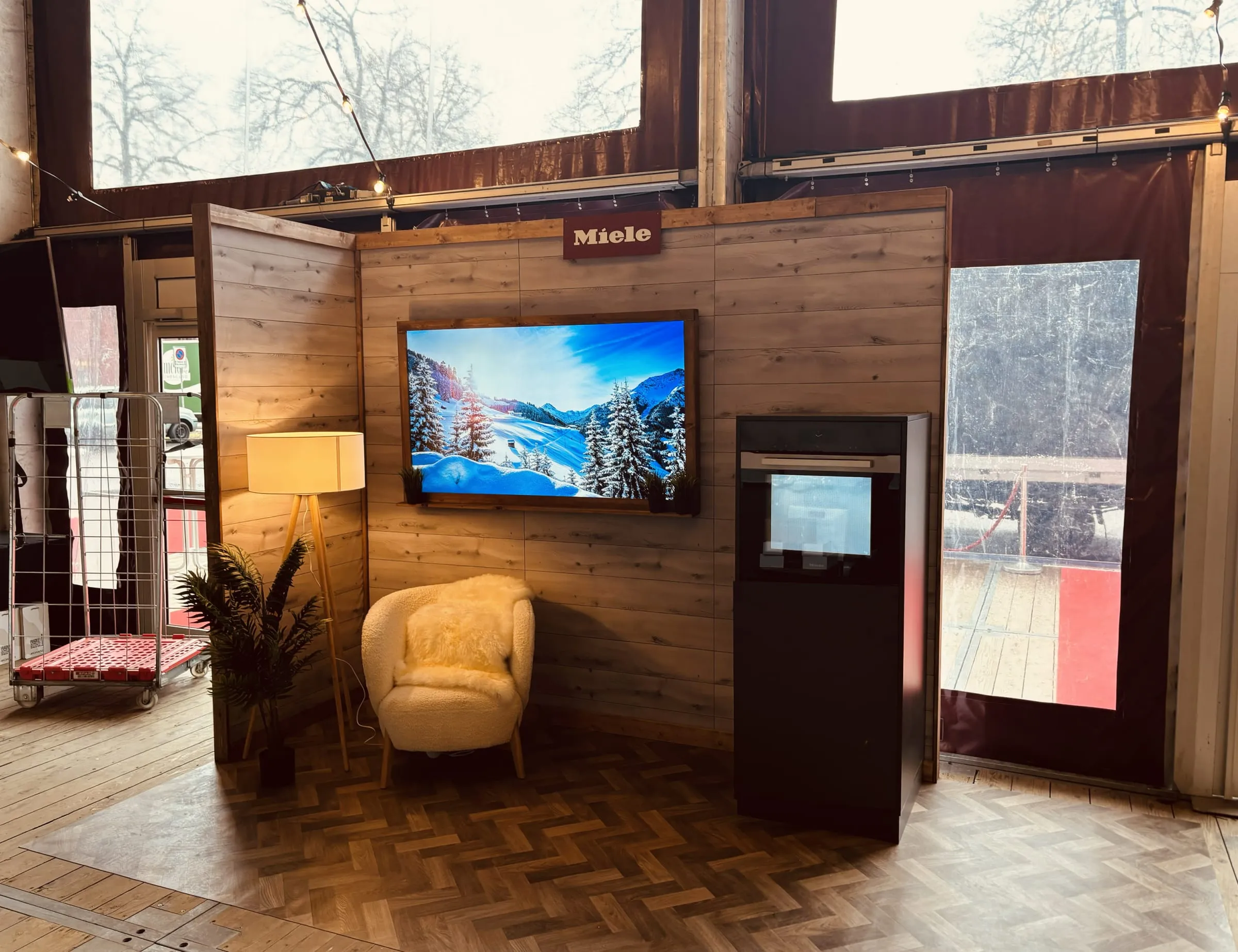 Frontansicht des Miele-Stands mit verschneiter Berglandschaft im Holzrahmen, Sessel und Stehlampe