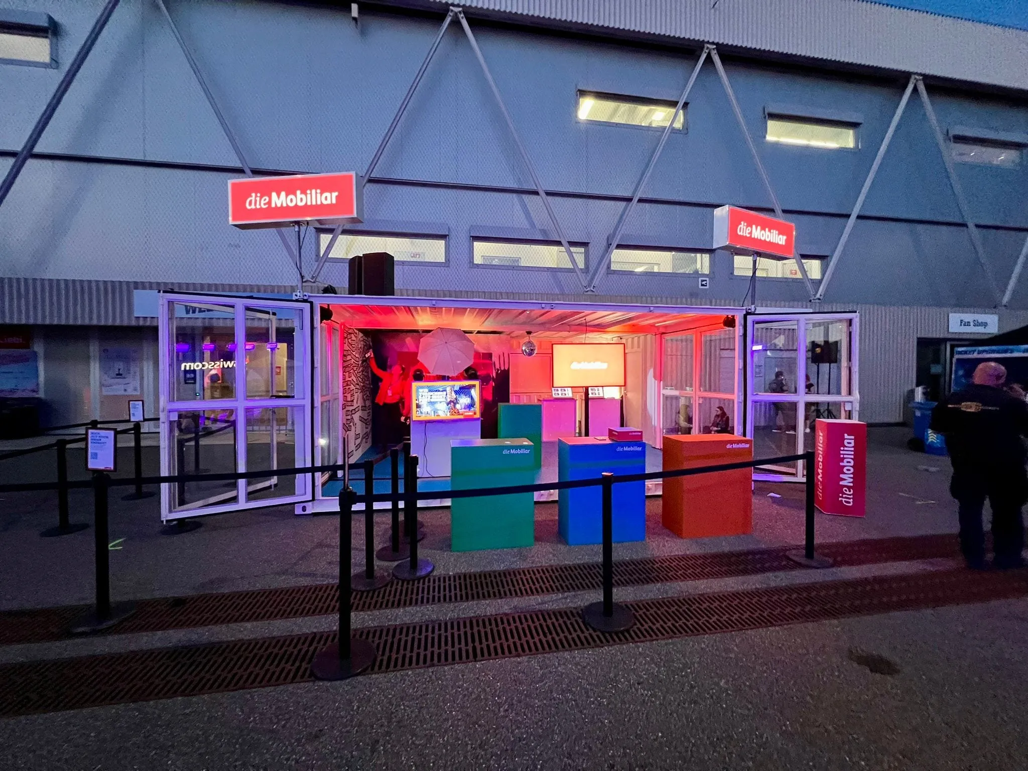 Container at night with red and blue interior light, word wall, screen and queue barriers in front