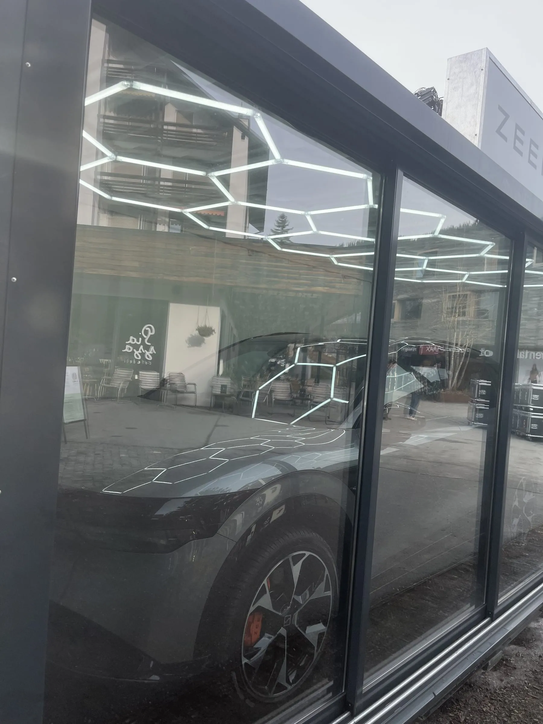 Blick durch die Glasfront auf die Front eines grauen Zeekr-Autos, Waben-LED-Decke und Gebäude spiegeln sich in der Scheibe