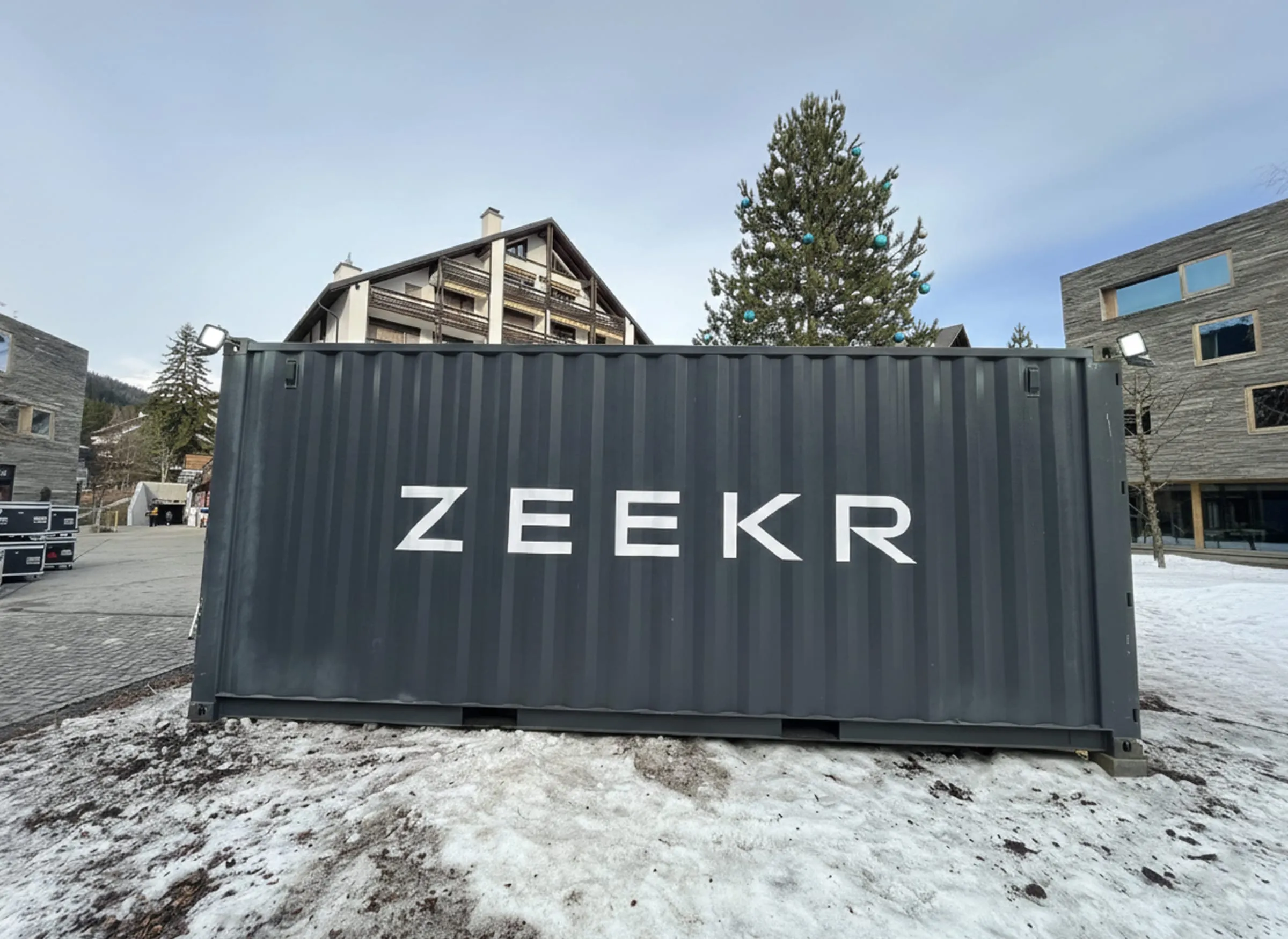 Rückseite des dunkelgrauen Containers mit grossem weissem «ZEEKR»-Schriftzug vor Chalet, Tanne und Steinfassade im Schnee