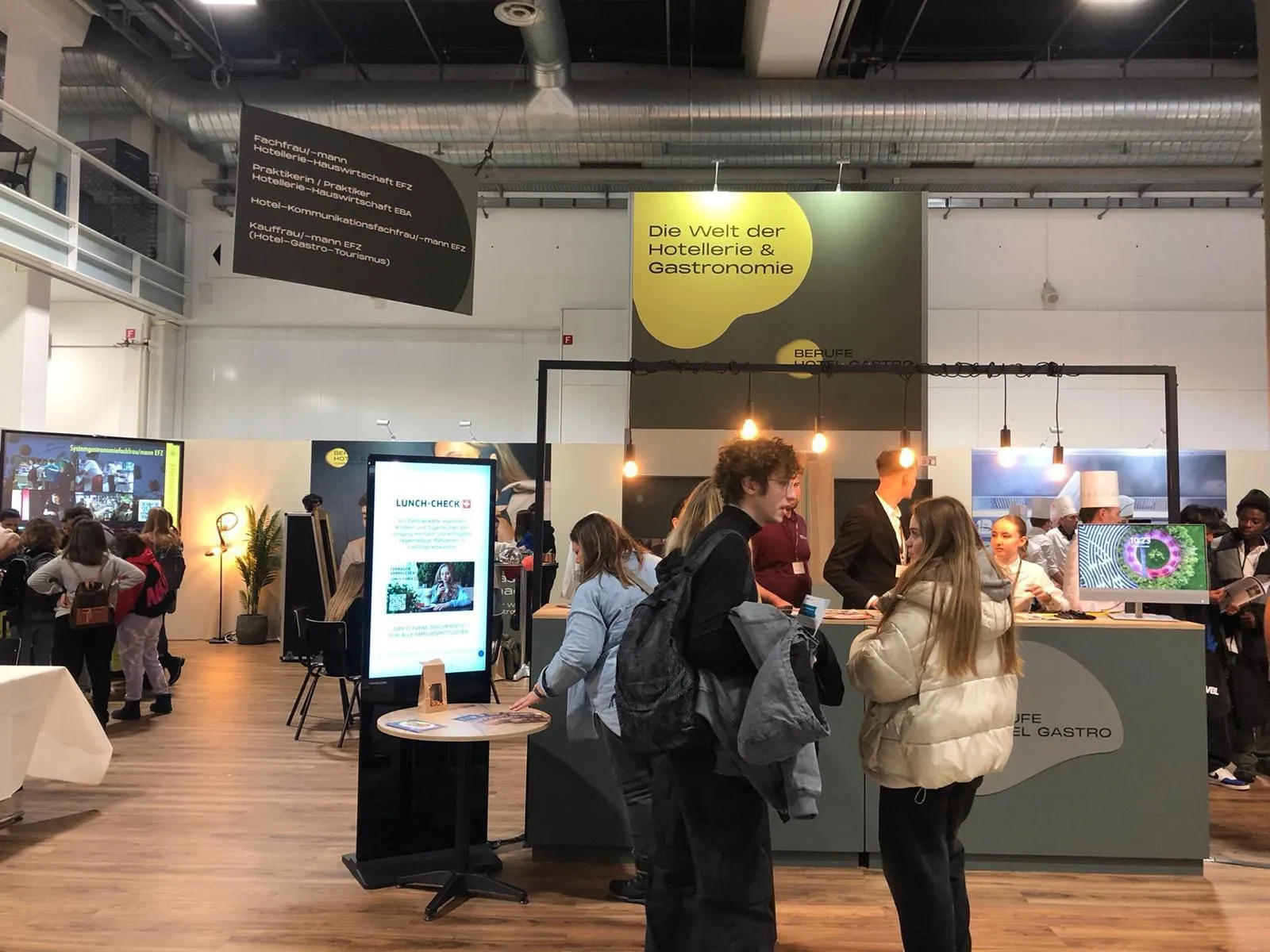 Jugendliche an der Theke des Stands mit Deckenschild «Die Welt der Hotellerie & Gastronomie» und Bildschirm