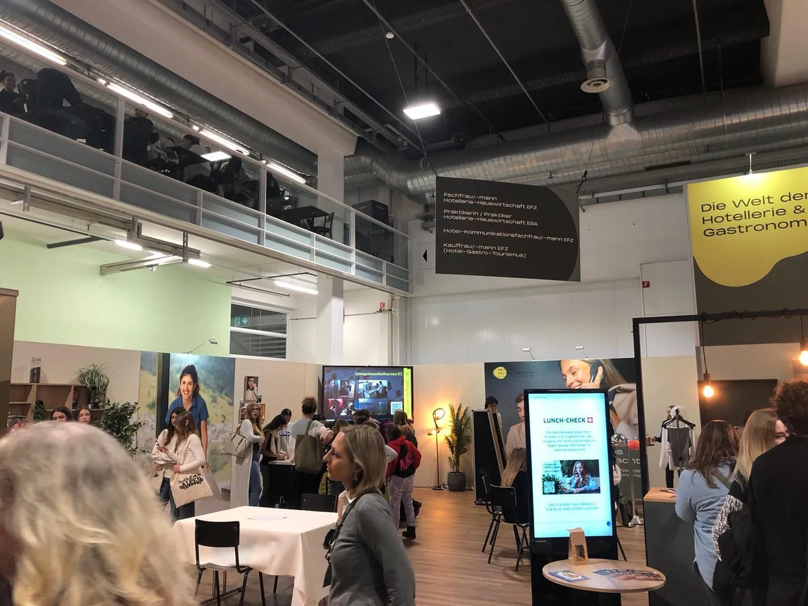 Besucher am Stand unter der Galerie, hängende Tafel mit Berufsbezeichnungen und Lunch-Check-Bildschirm