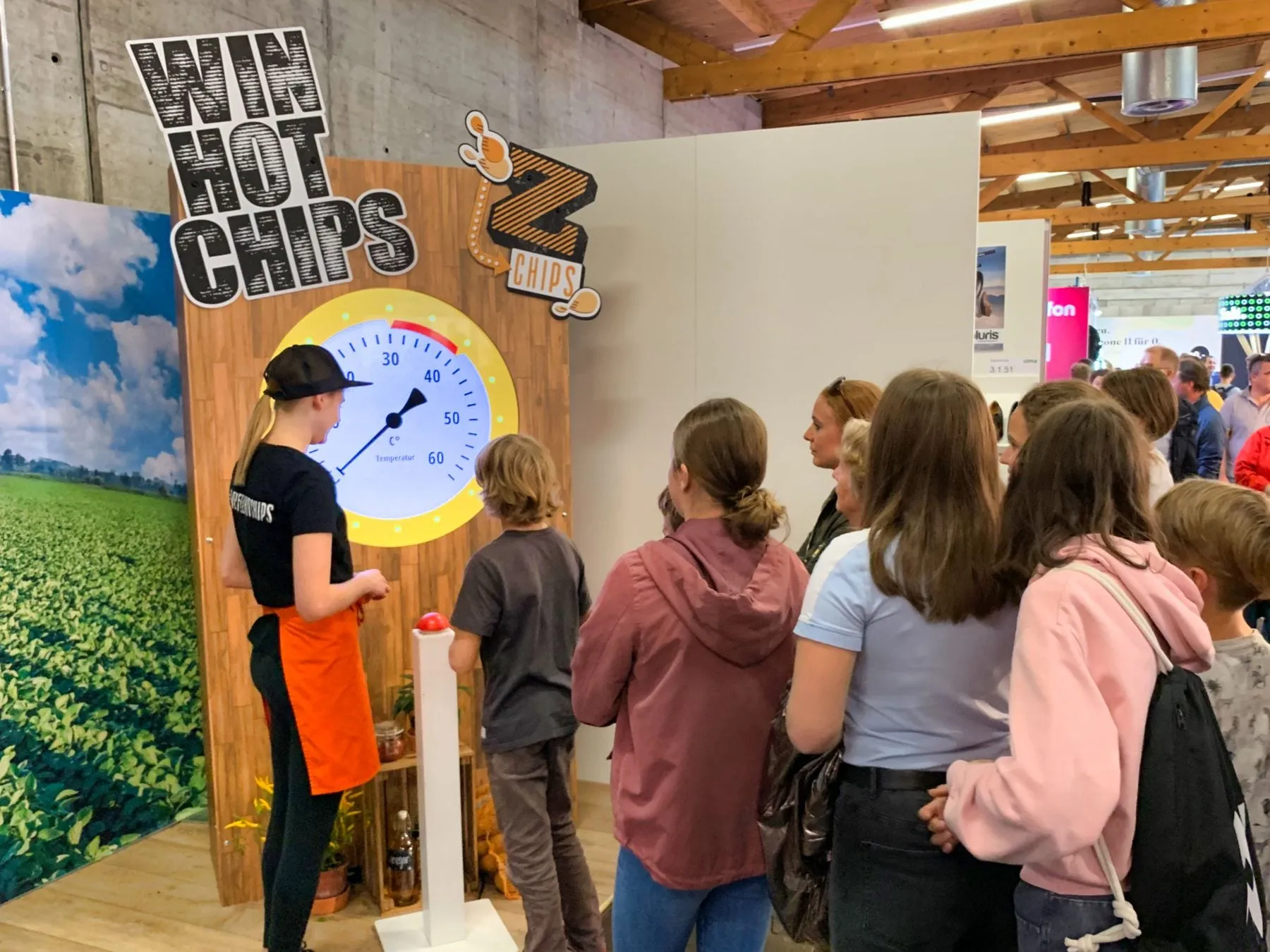 Wooden wall with «Win Hot Chips» lettering, a circular temperature display and a red buzzer, with children queuing