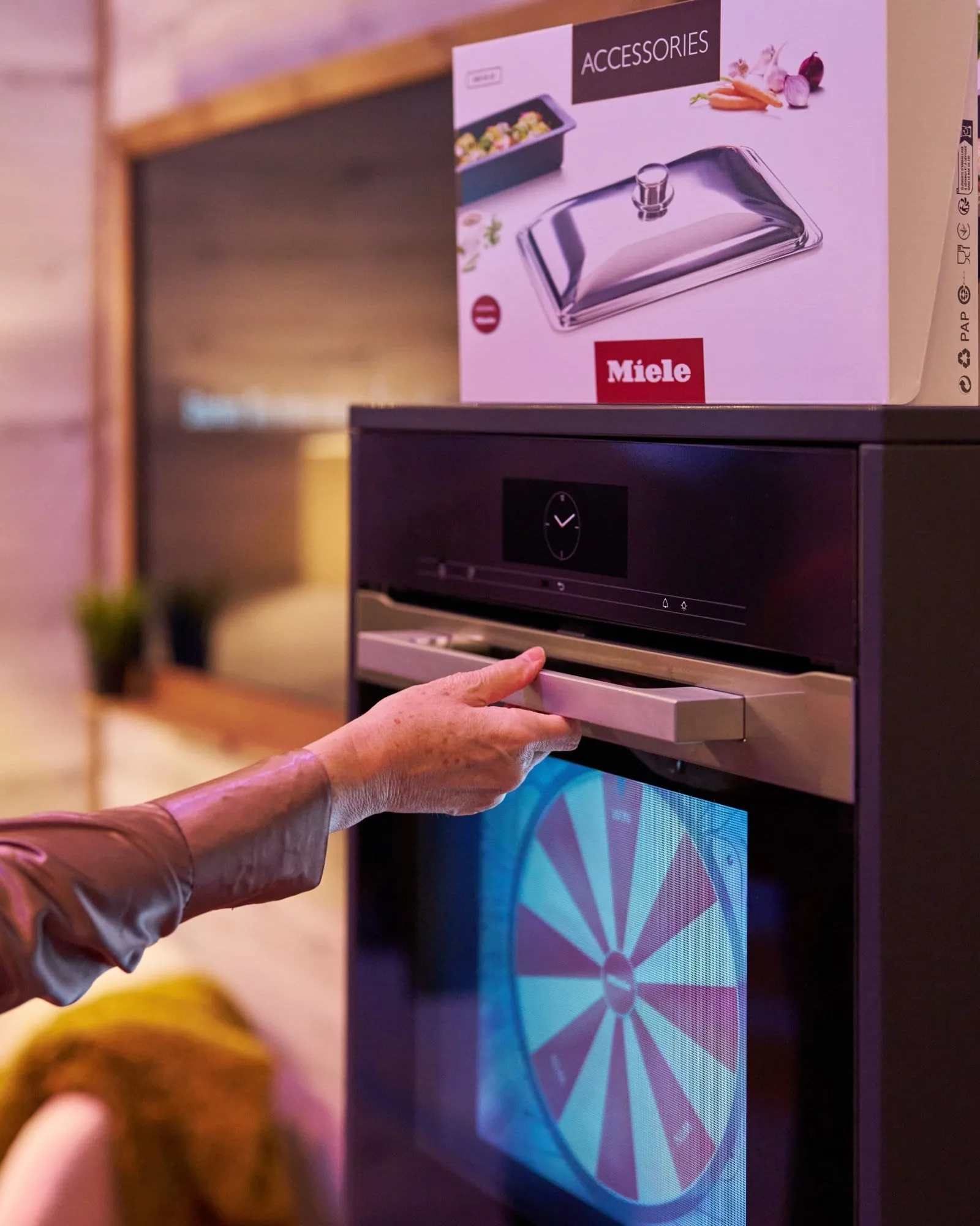 Hand greift an den Griff des Miele-Backofens, dahinter das Glücksrad auf dem Screen und eine Accessoires-Box