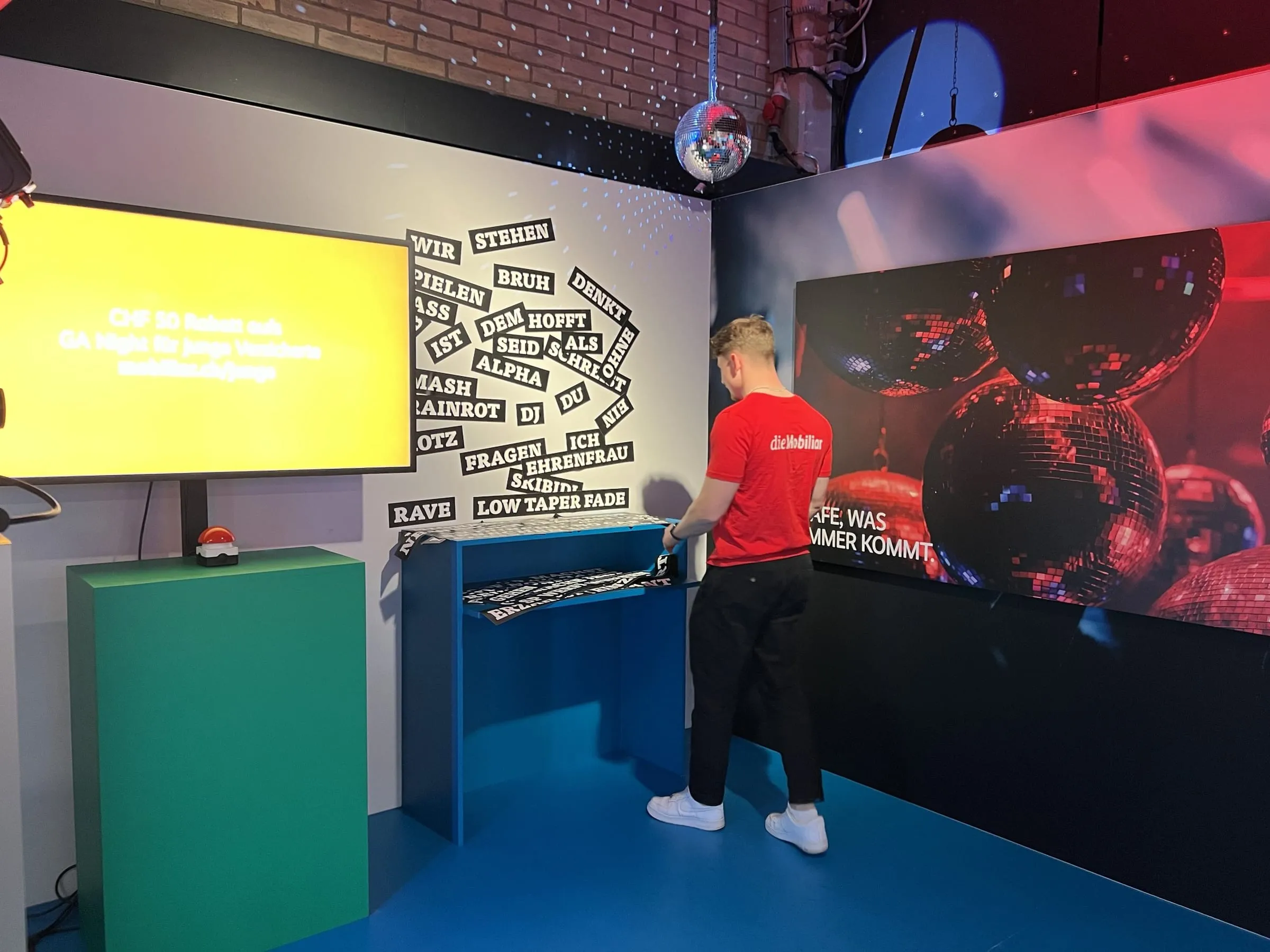 Promoter in a red Mobiliar shirt at the word wall with magnetic words such as «Bruh» and «Alpha», next to the red buzzer on a green pedestal