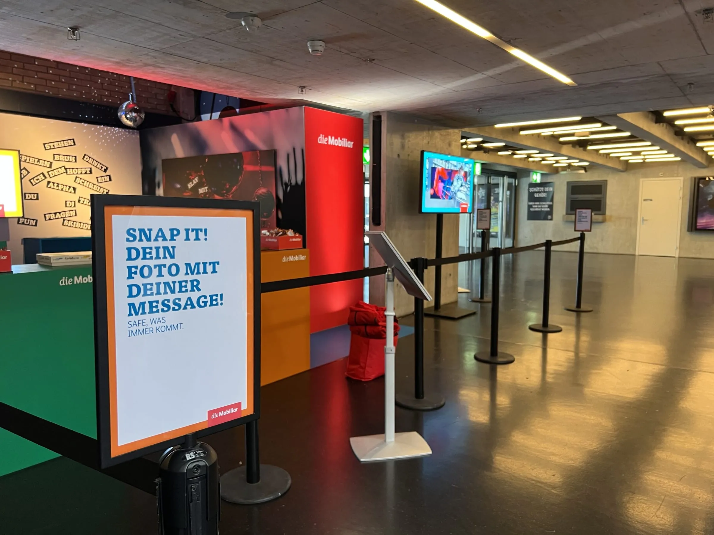 Sign «Snap it! Dein Foto mit deiner Message» in front of the stand, behind it queue barriers and the red Mobiliar cube